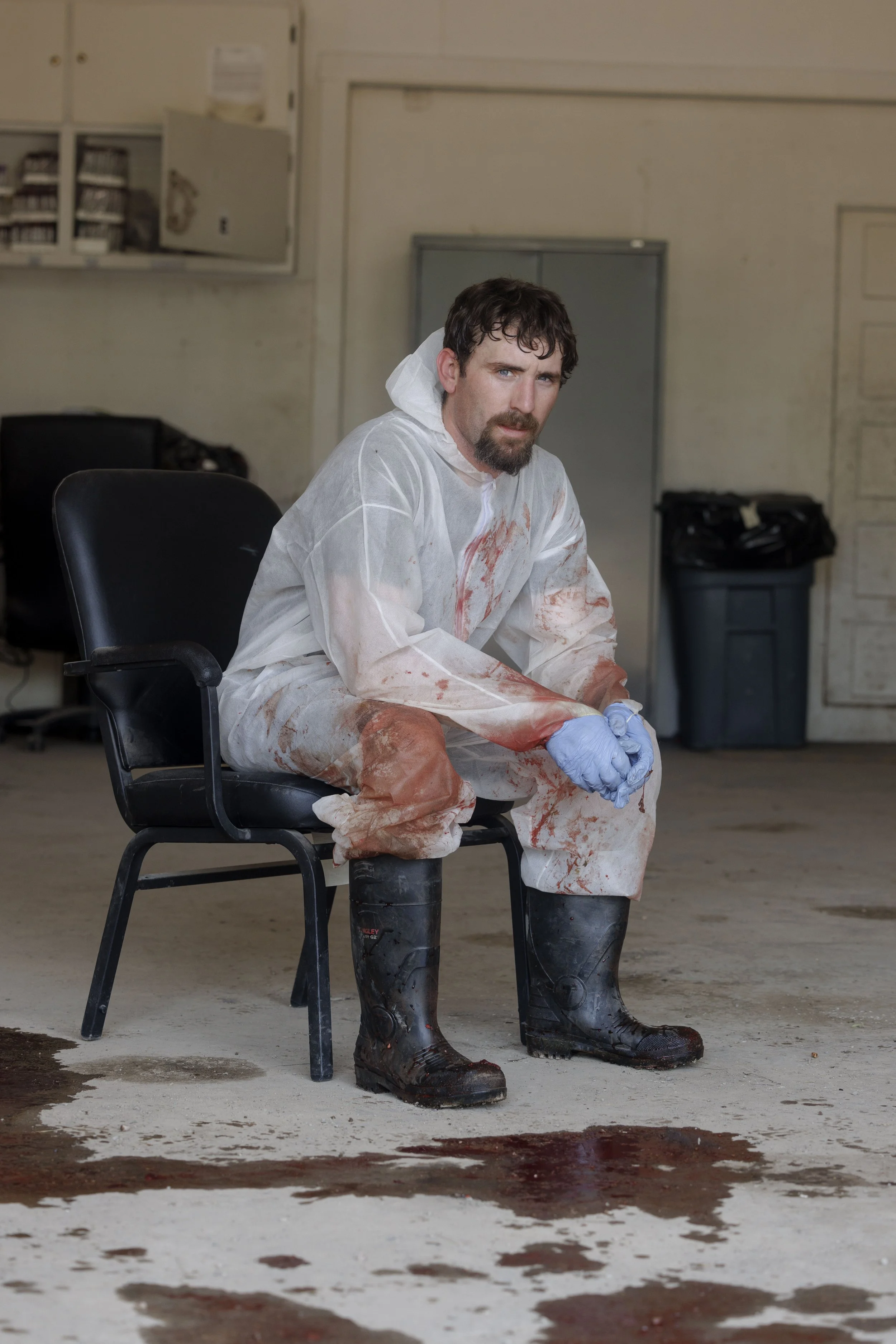 A man dressed in a bloodstained hazmat suit sitting on a black chair in an industrial or warehouse setting, with blood on his shoes and the floor beneath him.