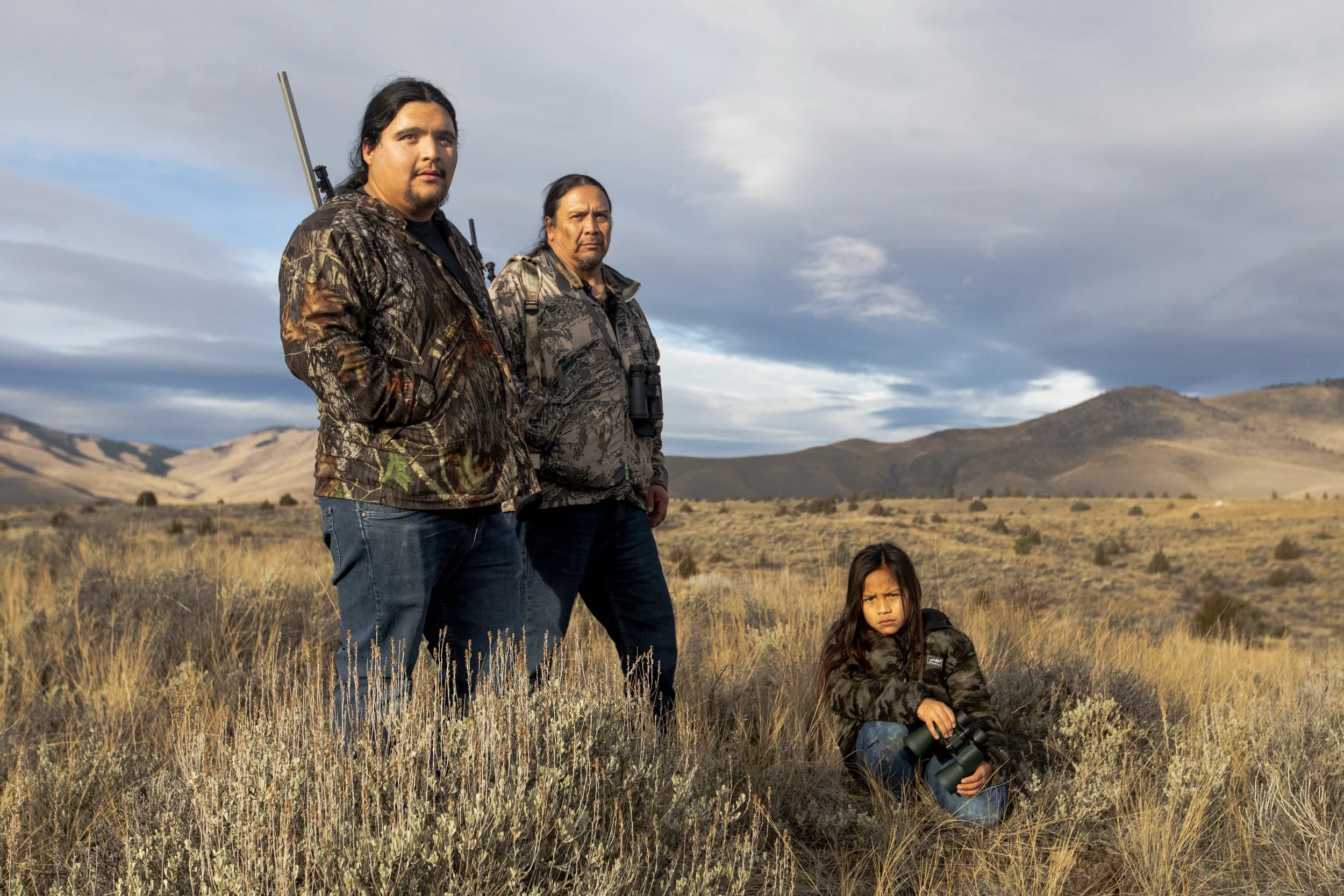 Three people, two adult men and one young girl, standing outdoors in a dry, grassy landscape with hills in the background. The men are wearing camouflage clothing and have serious expressions, with one holding a rifle and binoculars hanging around hi