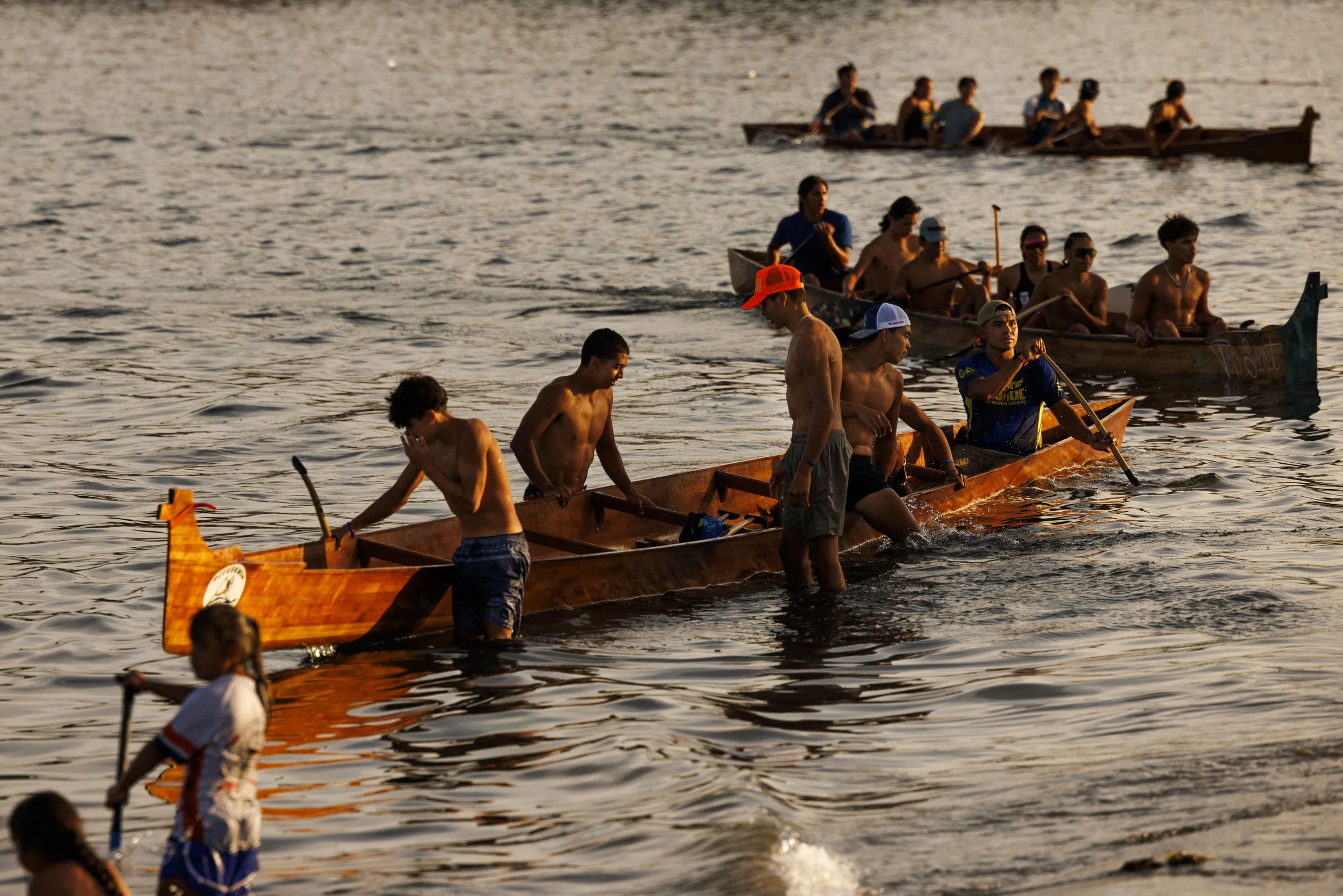 A group of young people in a boat and in the water during sunset, engaging in a dragon boat race or paddling activity on a calm body of water.
