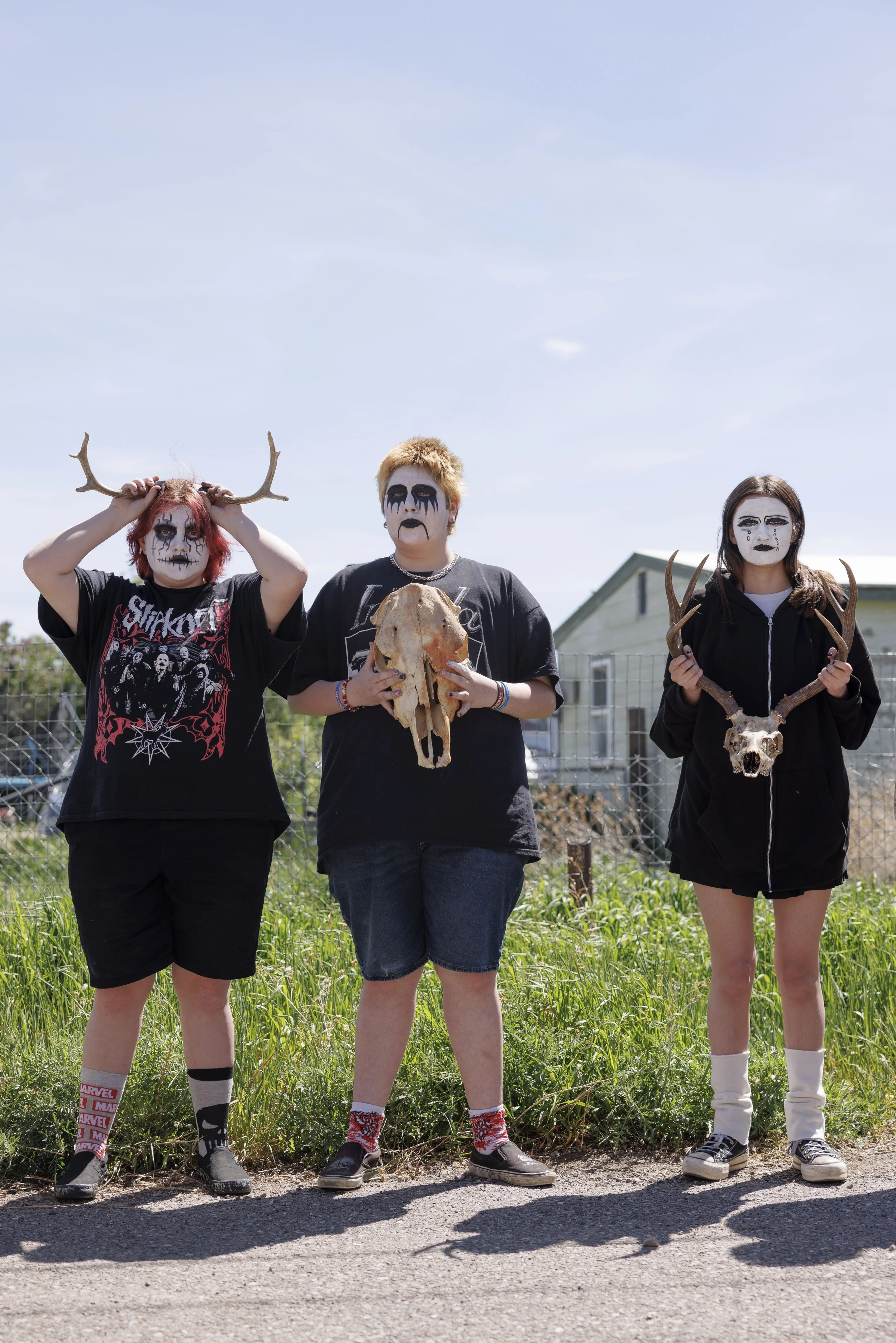 Three people dressed in Halloween costumes with face paint, holding animal skulls and antlers, standing outdoors on a sunny day with a house and fence in the background.