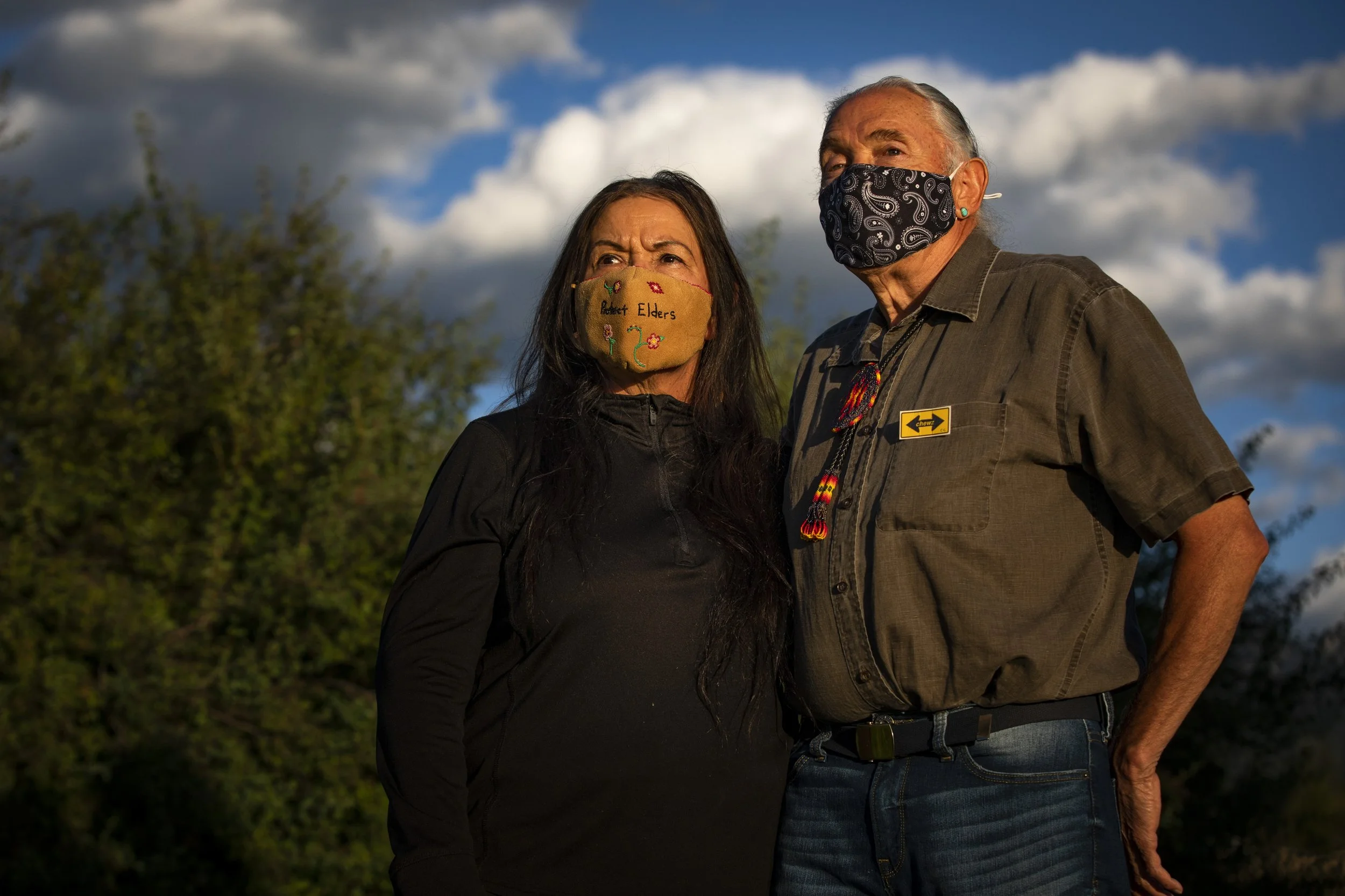 A man and a woman standing outdoors, wearing face masks, with a blue sky and clouds in the background.
