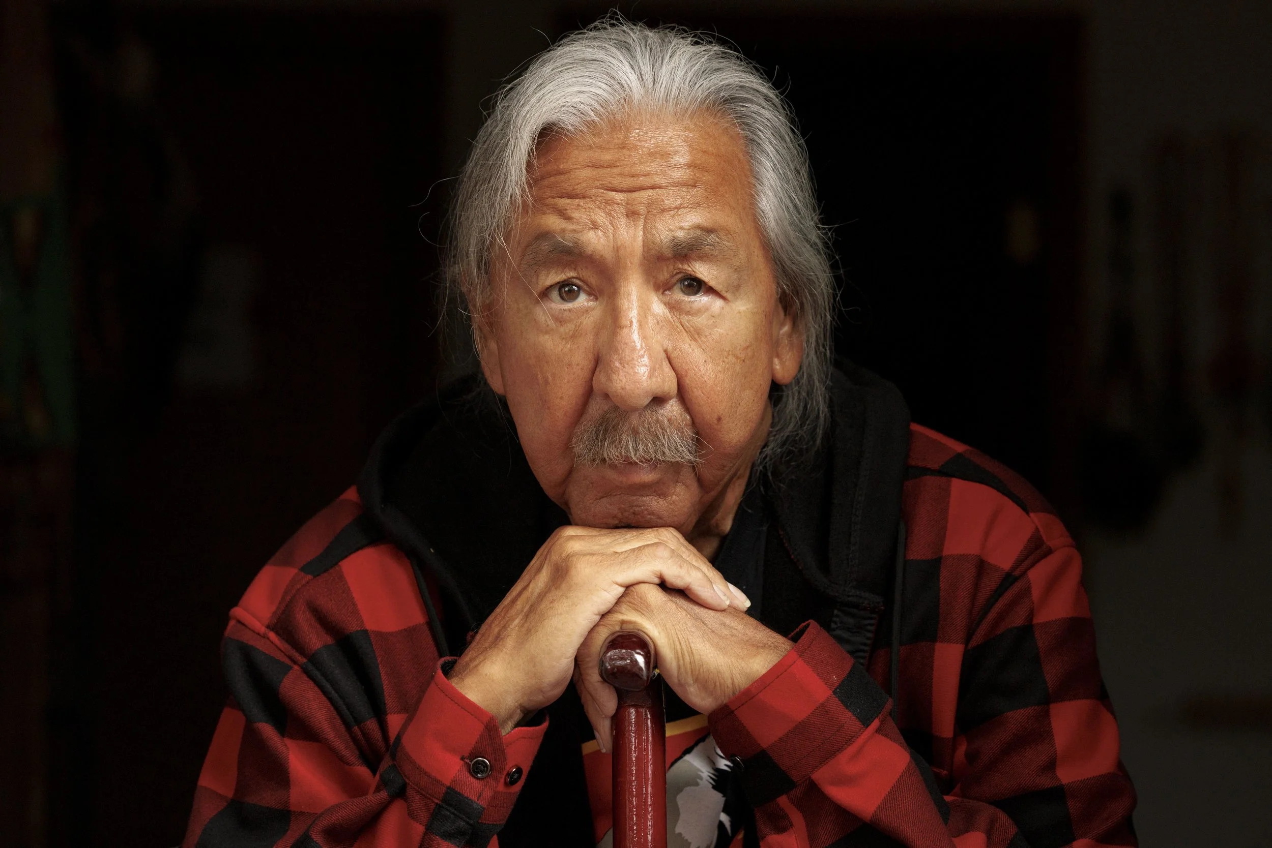 Portrait of an elderly man with gray hair, mustache, and beard, wearing a red plaid shirt, resting his chin on clasped hands and holding a cane, with a dark background.