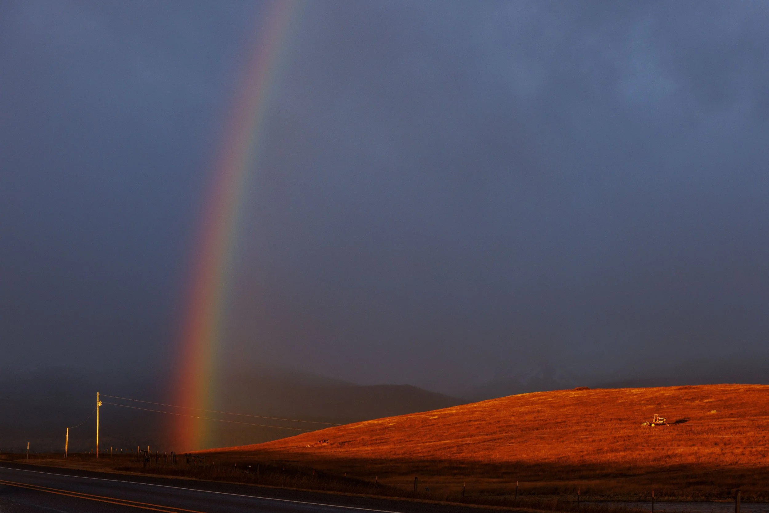 A rainbow with vibrant colors appears in a dark sky over a rural landscape with rolling hills and a road, illuminated by warm sunlight.
