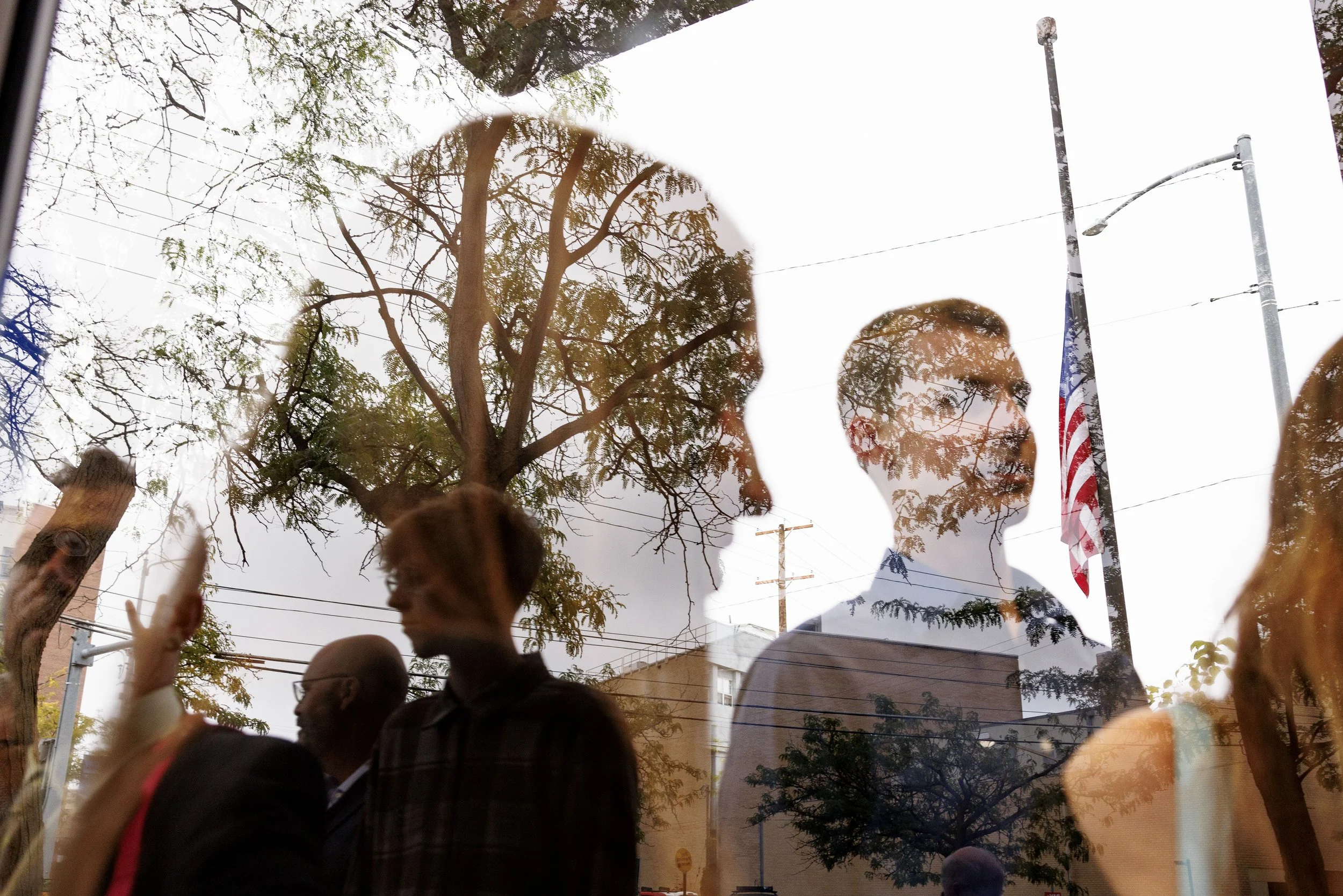 A reflection on glass showing a group of people, trees, power lines, streetlights, and an American flag.