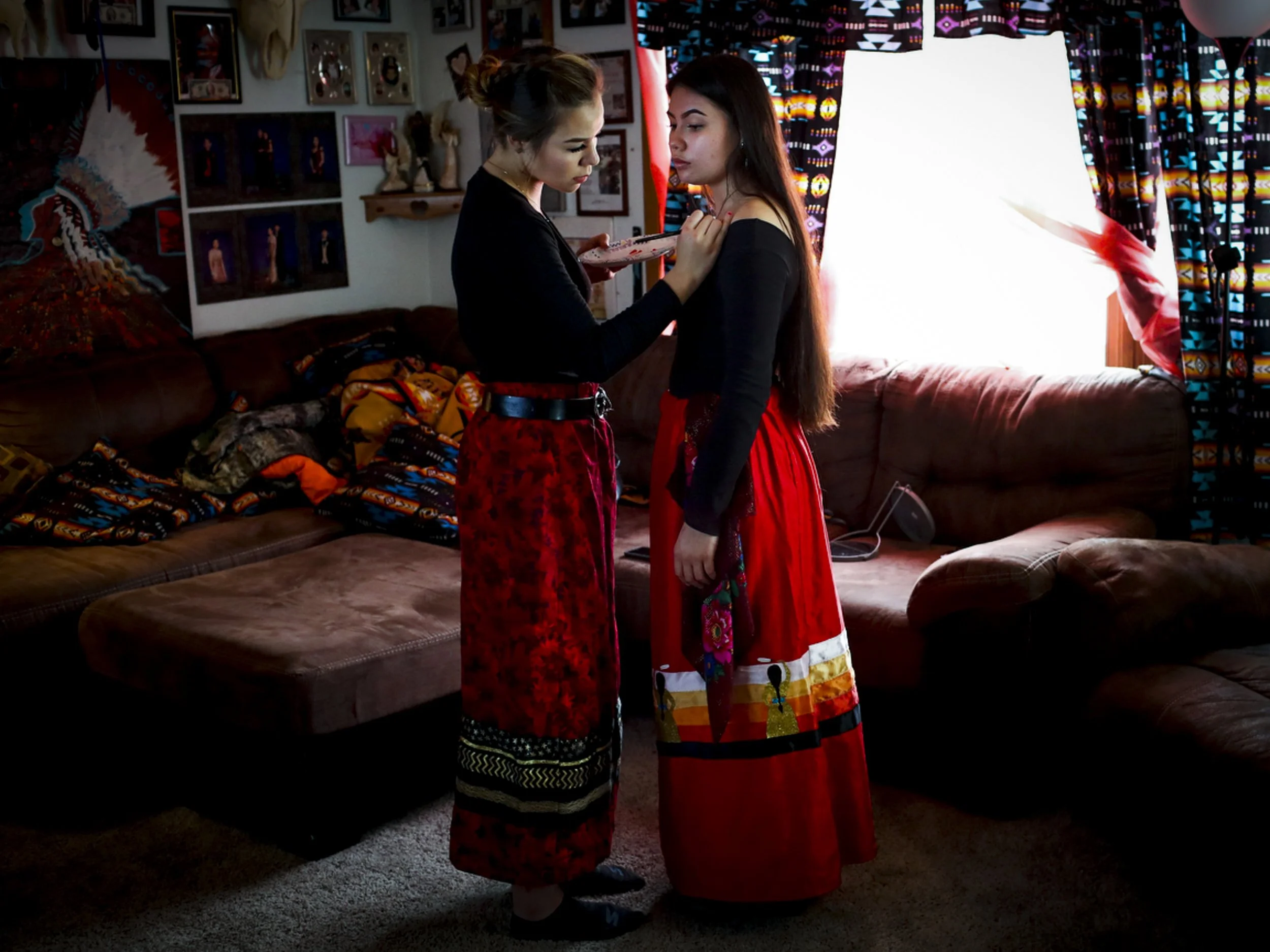 Two young women in colorful traditional Mexican dresses are standing close together in a cozy, decorated living room. One woman is applying makeup or face paint to the other.