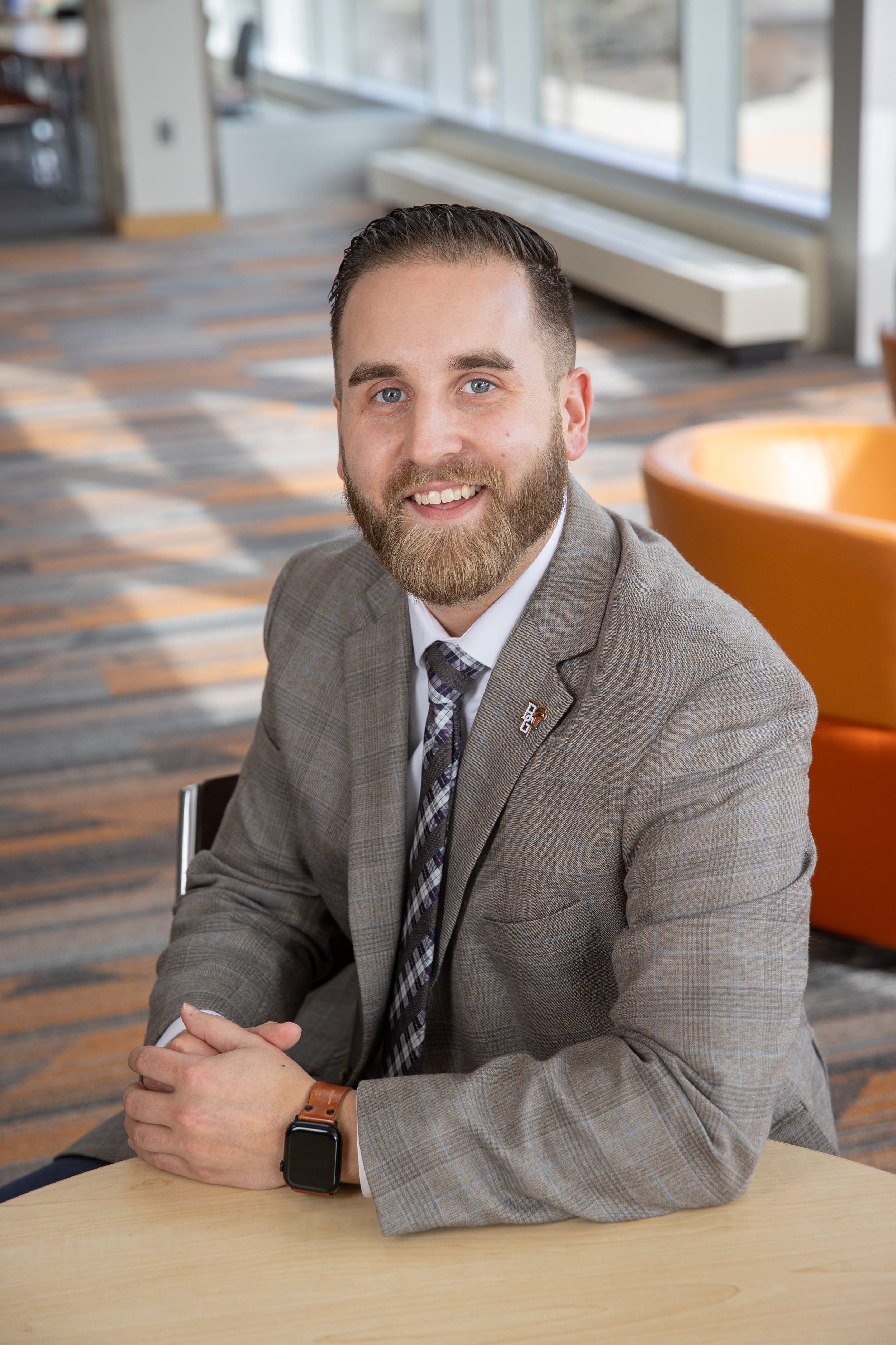 A man with a beard and light blue eyes, wearing a gray plaid suit, white shirt, and a patterned tie, is sitting at a wooden table with his hands clasped. He is in an indoor setting with large windows, colorful carpet, and orange chairs.