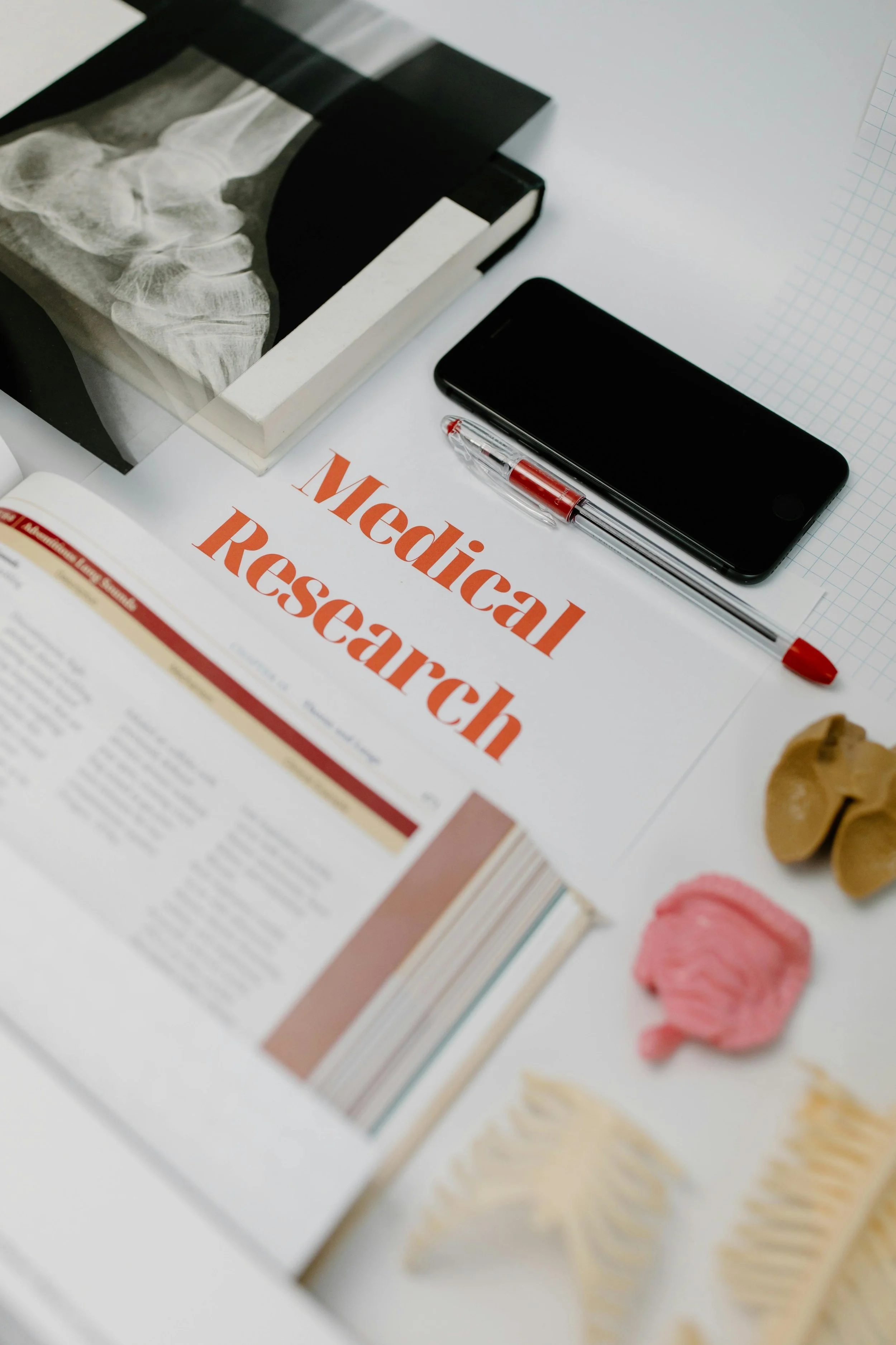 Study desk with a book titled "Medical Research," a smartphone, a tube pen, a graph paper on a white surface, and some crumpled paper and snacks nearby.
