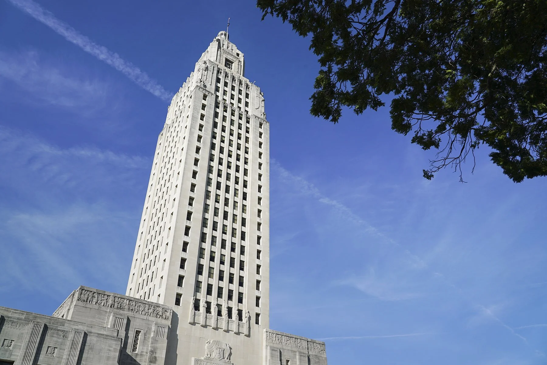 Baton Rouge state capitol building, a tall white skyscraper with art deco architecture, seen against a bright blue sky with a tree's branches and leaves in the upper right corner.
