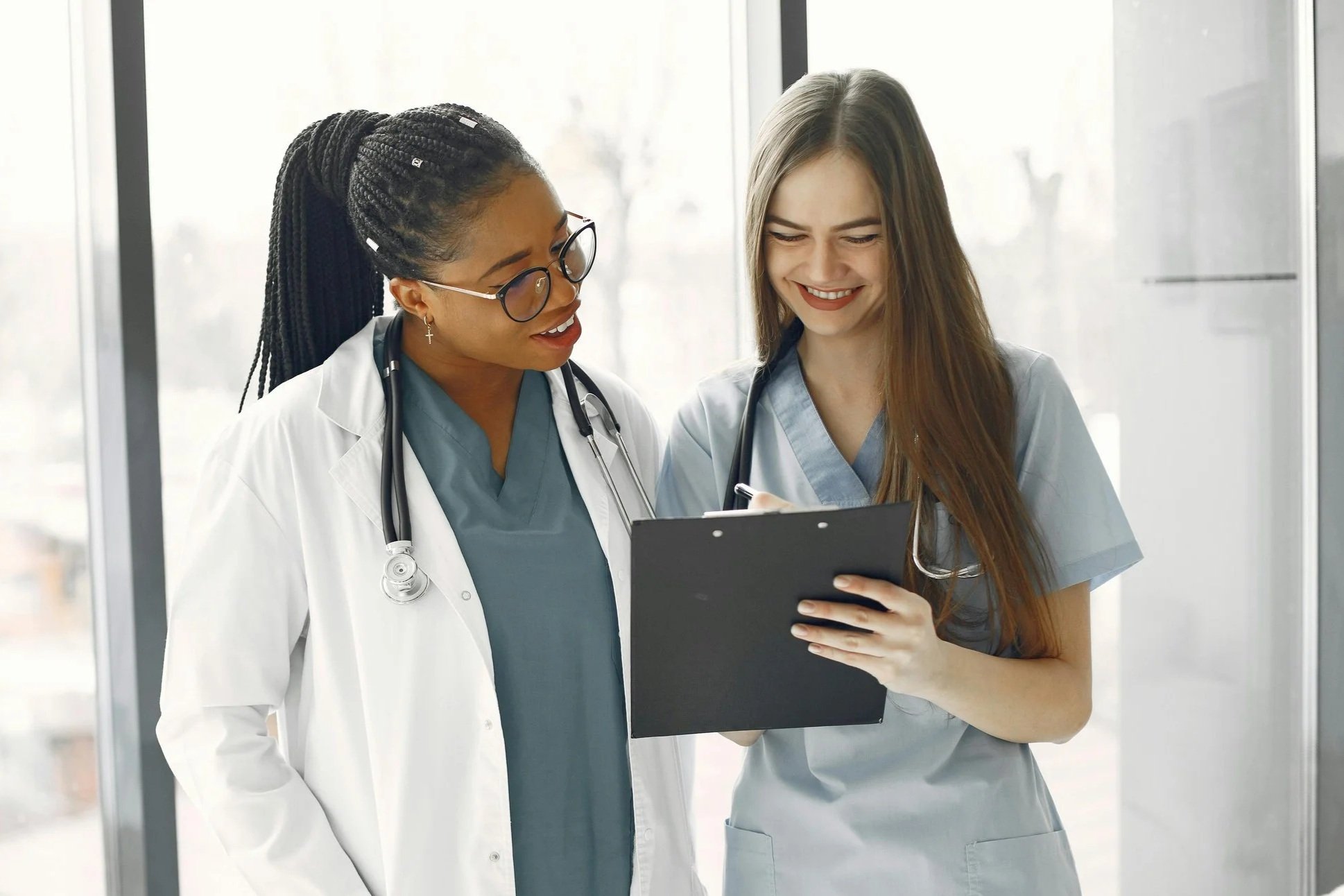 Two female healthcare professionals, one with glasses and a stethoscope, and the other with long brown hair and a stethoscope, looking at a clipboard and smiling in a medical facility.