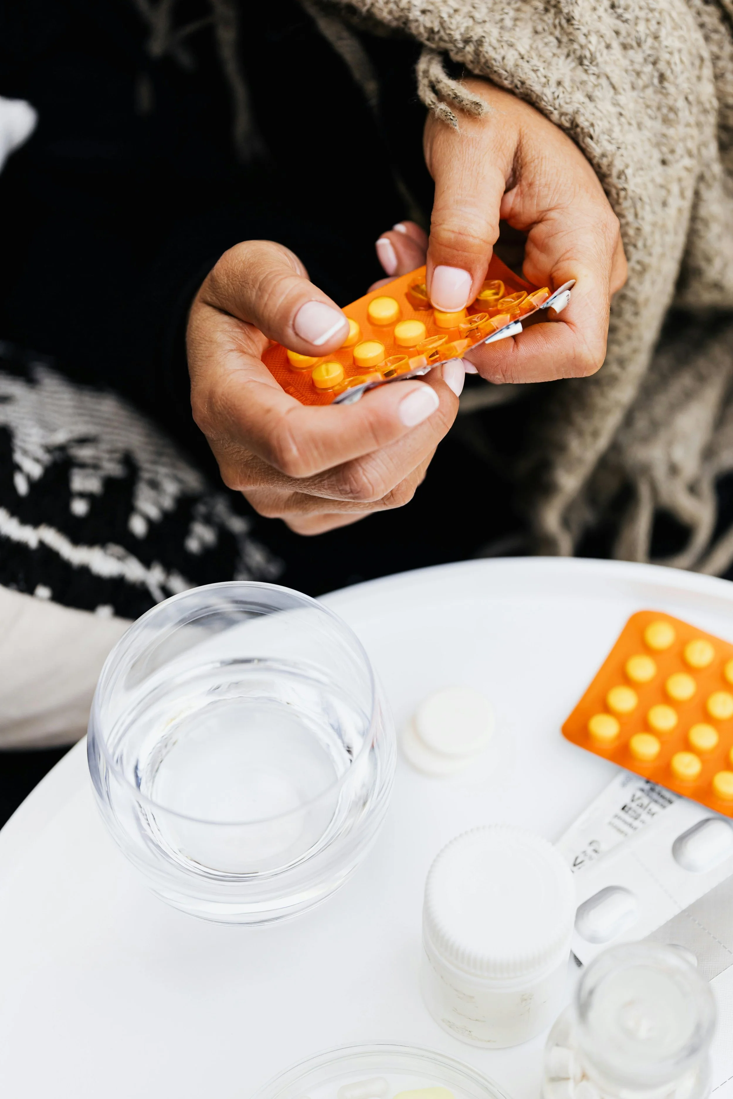 A person's hands holding an orange blister pack of yellow and orange pills, with a glass of water and white medication bottles on a white table.
