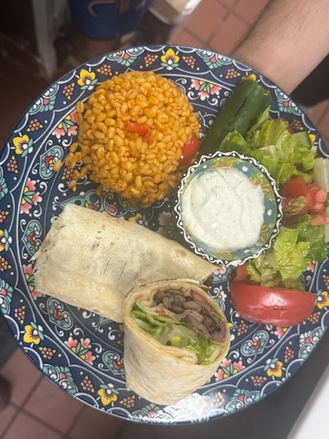 A decorative plate holding a serving of seasoned rice, a beef and lettuce wrap, a vegetable spring roll, a small bowl of creamy dipping sauce, and a side salad with tomato, cucumber, and lettuce.