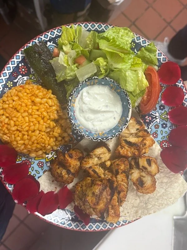 Plate of grilled chicken, rice, salad with lettuce, tomato, and radishes, sliced cucumber, and a small bowl of creamy dressing, garnished with red rose petals.