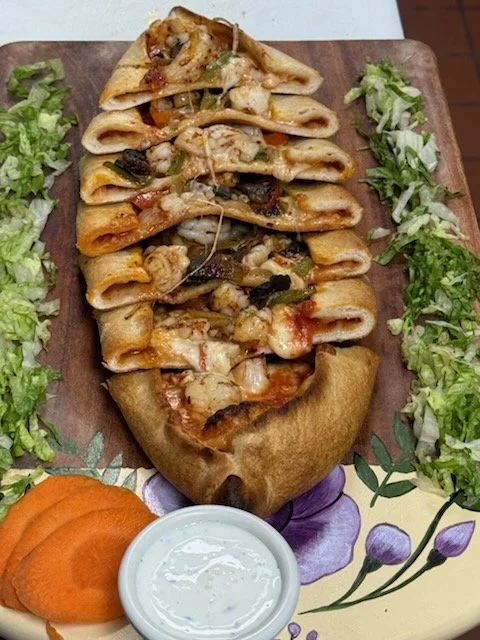 A pizza shaped like a boat with assorted toppings, surrounded by shredded lettuce on a floral plate, with sliced carrots and a small dish of white dipping sauce.