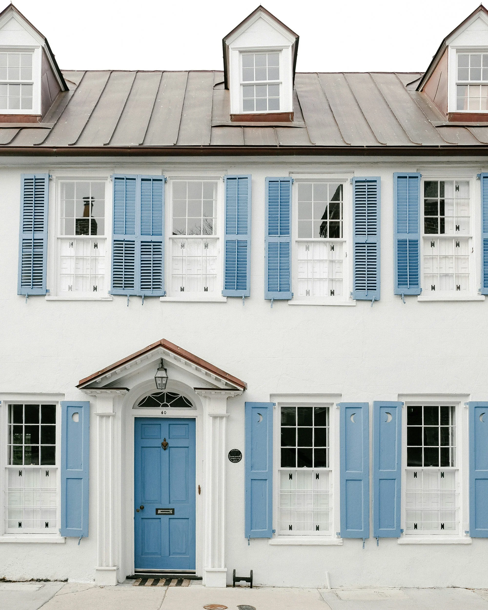 Two-story white house with blue shutters and a blue front door, featuring two dormer windows on the roof and a small front porch with a lantern light.