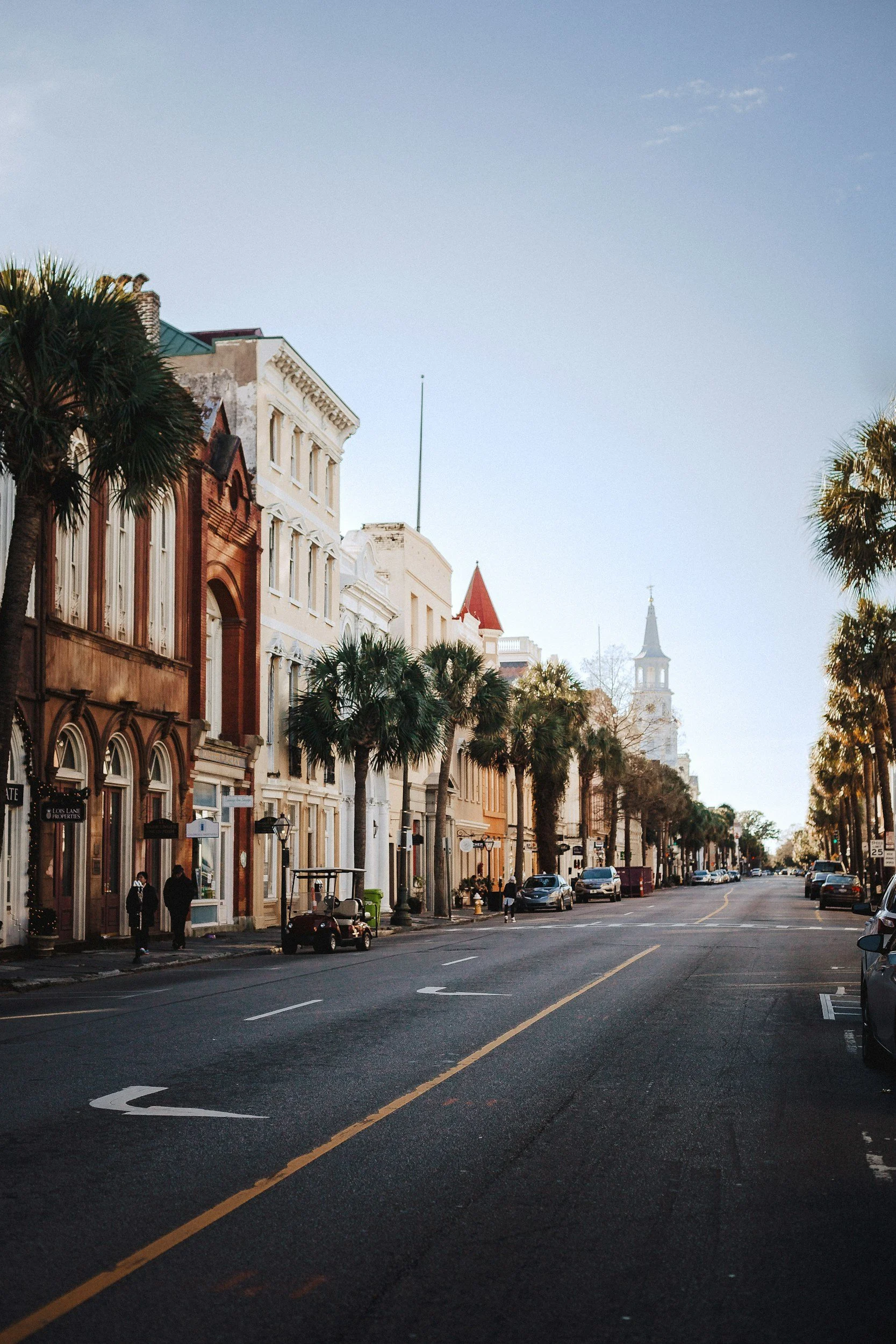 A city street with historic buildings on the left side, palm trees lining the sidewalk, and parked cars along the curb. The sky is clear with a few clouds, and there's a church tower visible in the background.