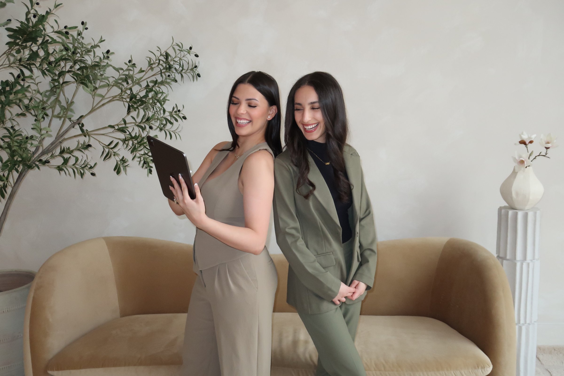 Two women are standing side by side in a living room. The woman on the left is holding a tablet, while the woman on the right is standing with her hands clasped in front of her. Both women are smiling and appear to be looking at the tablet. There is a large green plant on the left and a white vase with white flowers on the right, with a beige sofa behind them.