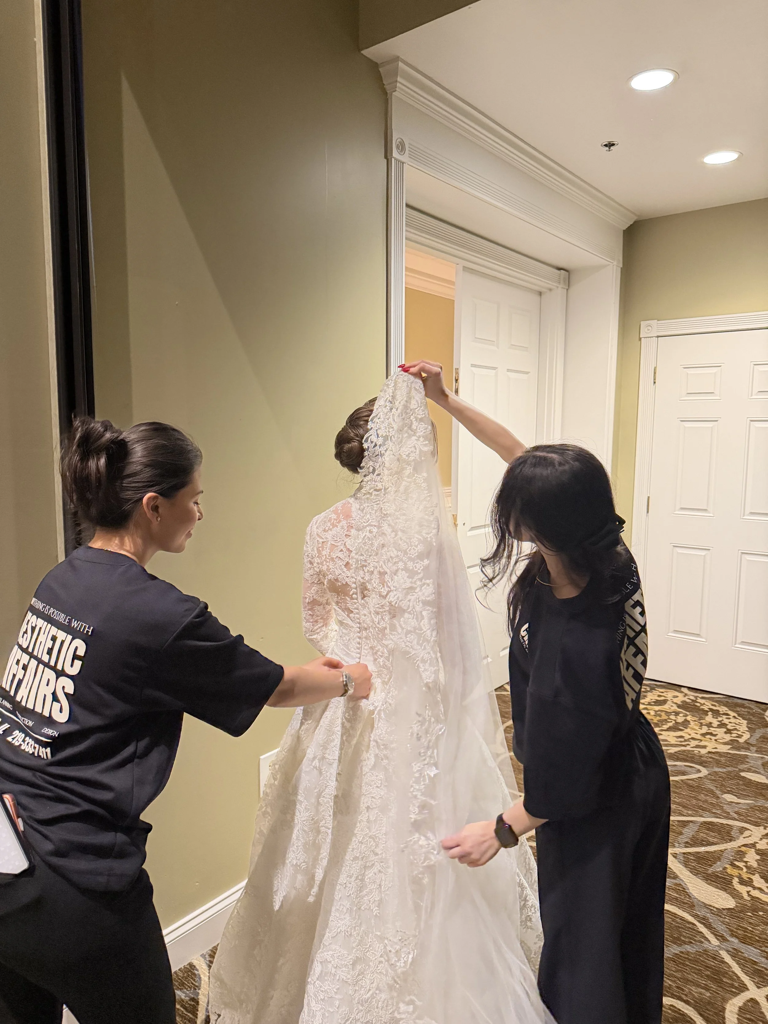 Three women are dressing a bride in a white lace wedding gown in an indoor setting, preparing her for her wedding.