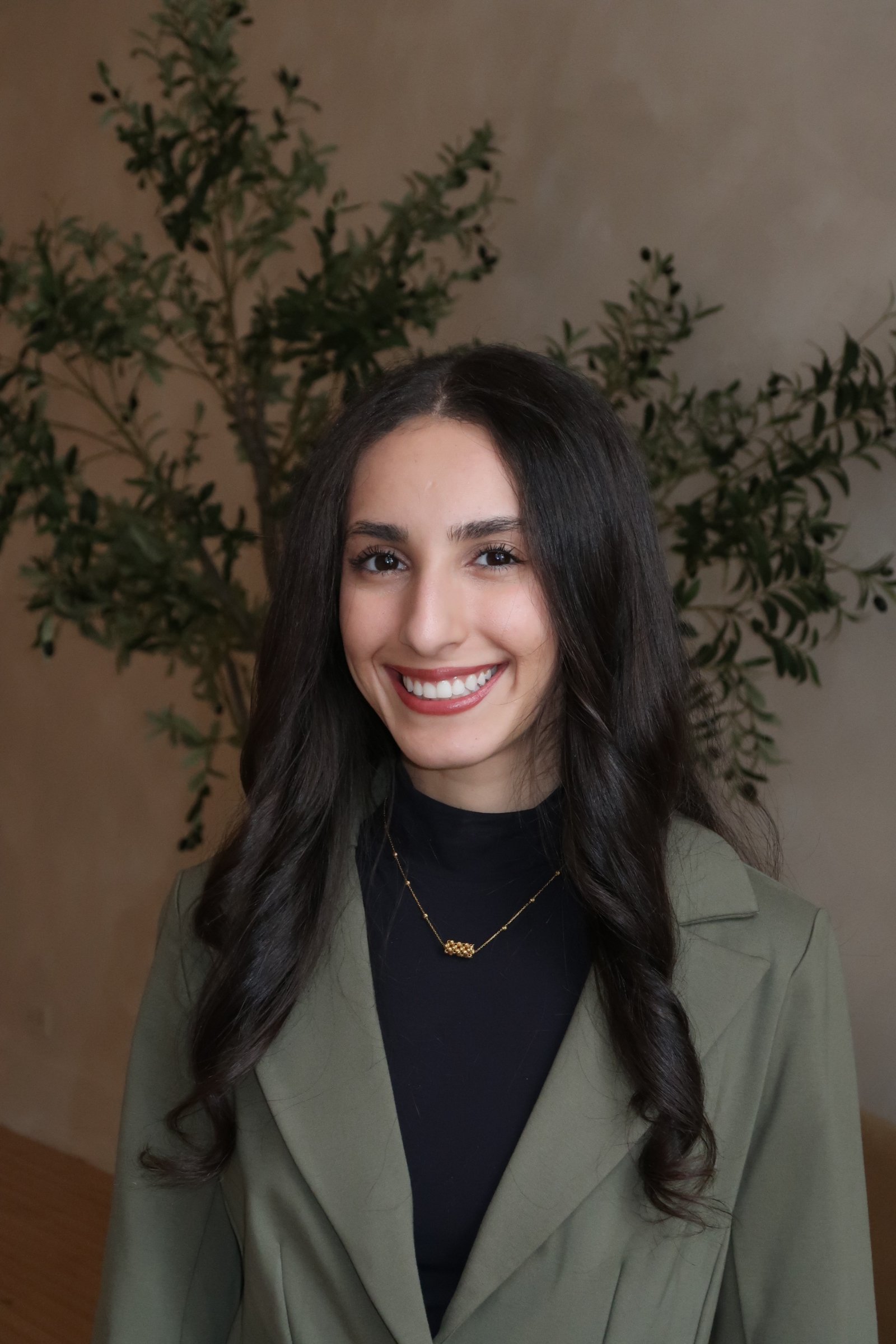 Young woman with long dark wavy hair smiling, wearing a black top, layered gold necklaces with a charm, and a green blazer, standing in front of a beige wall with a leafy green plant.
