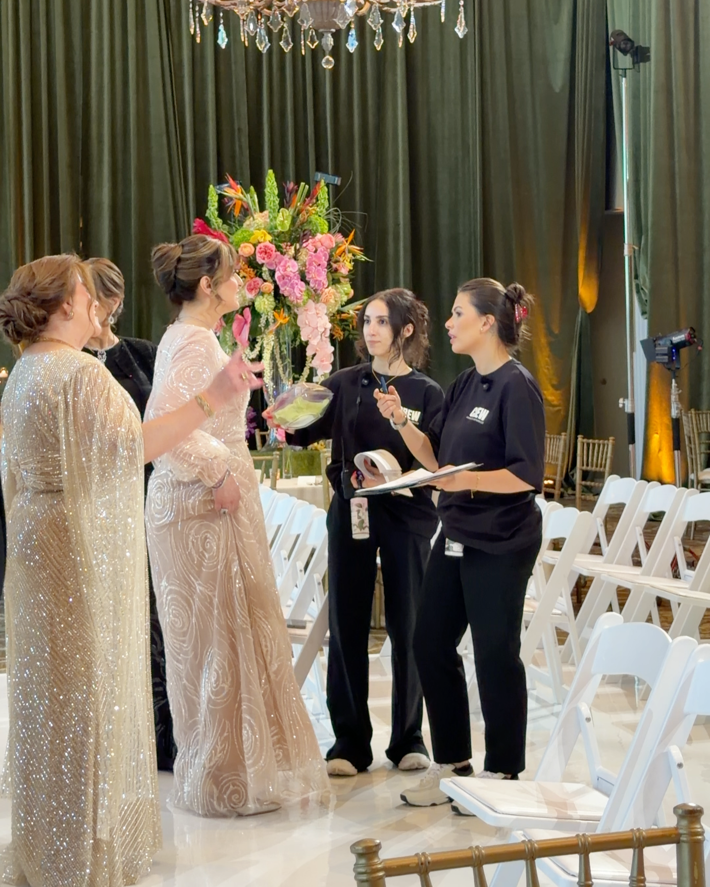 Four women, two in elegant gold dresses and two in casual black shirts, are having a conversation in a decorated event space with a large floral arrangement in the background. There are white chairs arranged in rows and a chandelier hanging above.