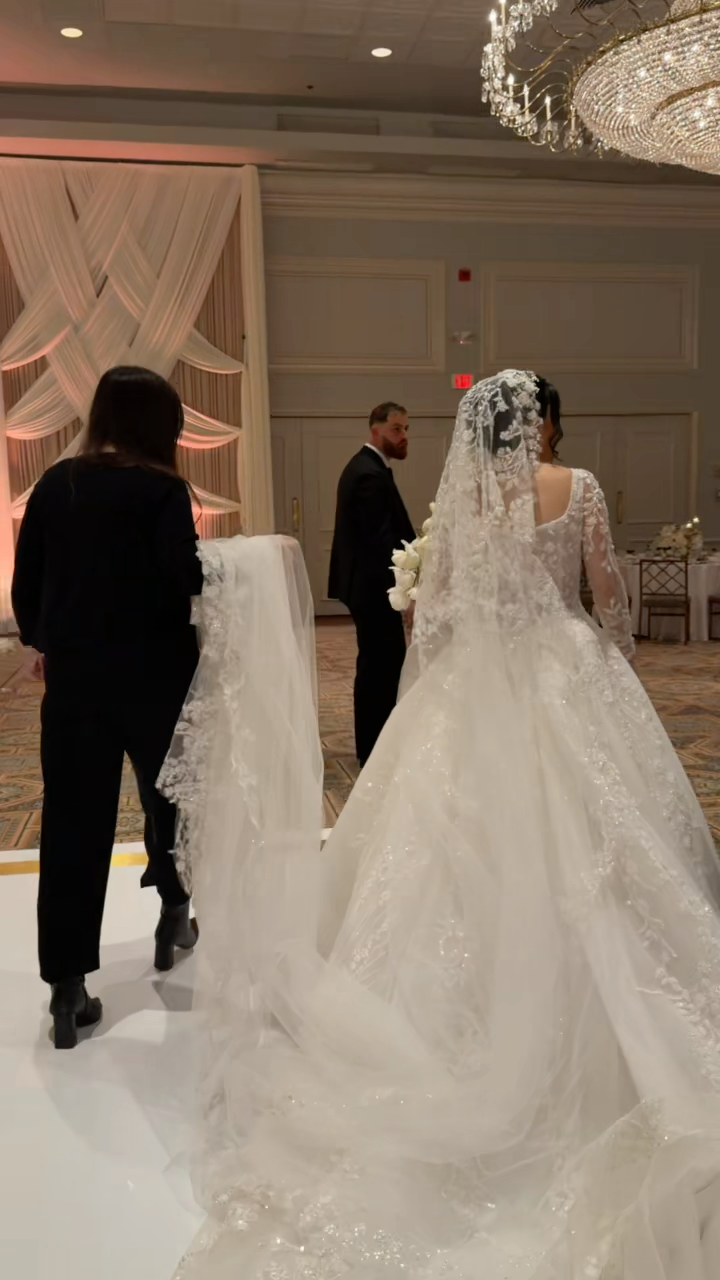 A bride in a white wedding gown and veil holding a bouquet, standing in a decorated wedding venue with a chandelier and draped fabric backdrop. Two people are adjusting her dress.