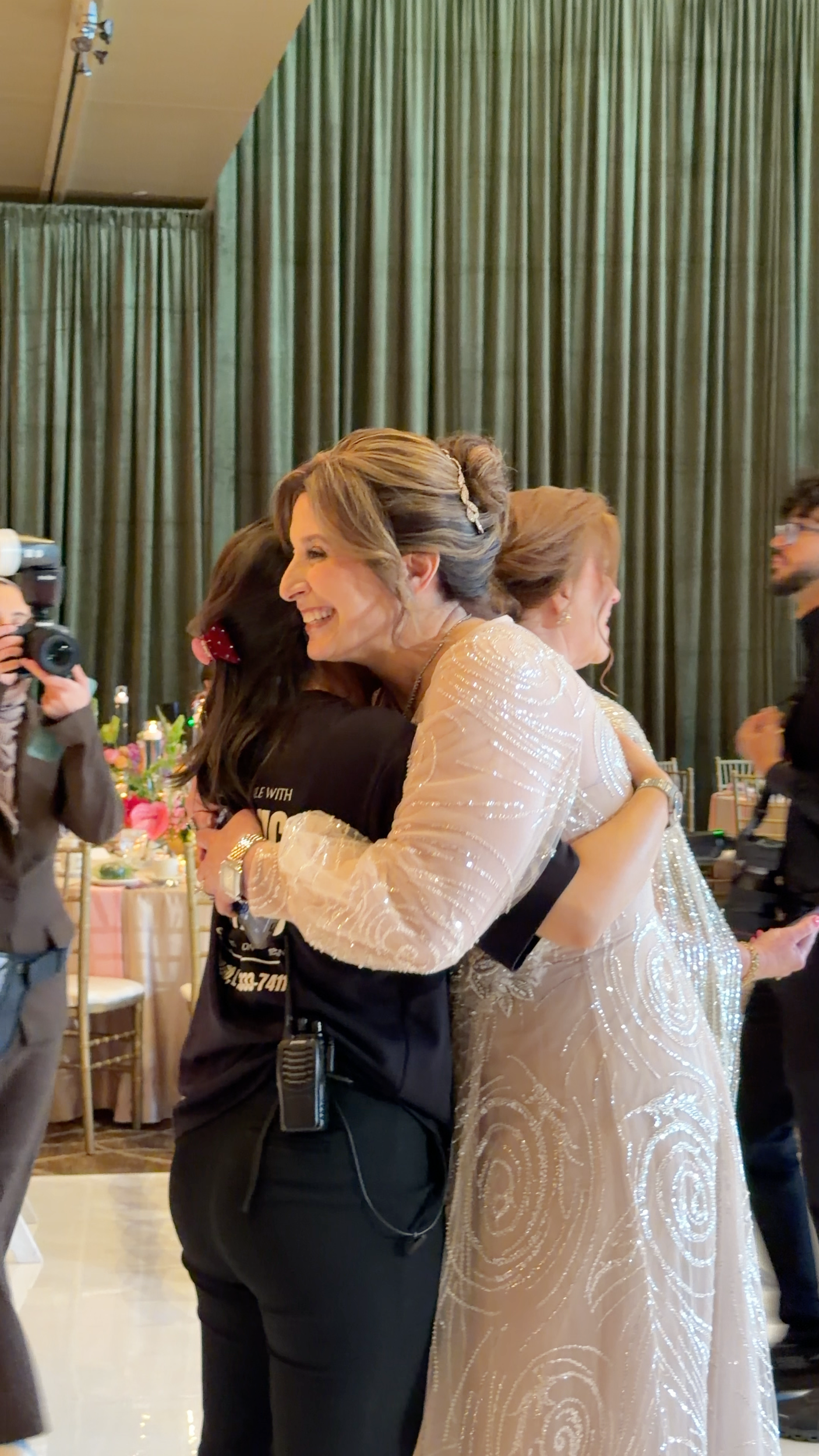 A woman in an elegant, beaded white dress hugging a younger woman at a celebration event with green curtains and decorated tables in the background.