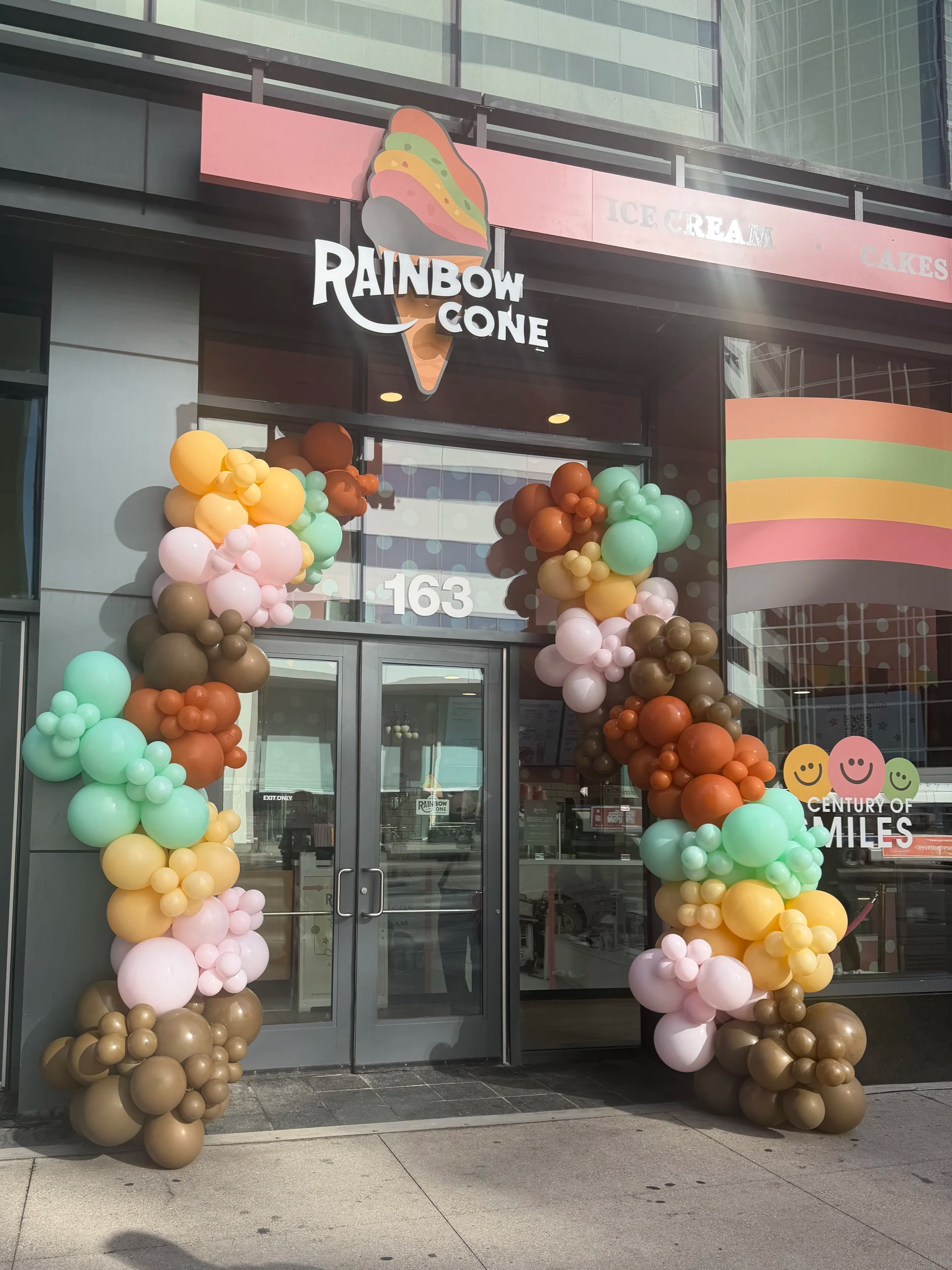 Entrance of Rainbow Cone, decorated with colorful pastel balloons forming two columns on each side of the door and a rainbow-colored banner on the window. The Rainbow Cone logo, featuring a cartoon ice cream cone with rainbow swirls, is above the door. The store is located at 163, with large glass windows showing the interior.