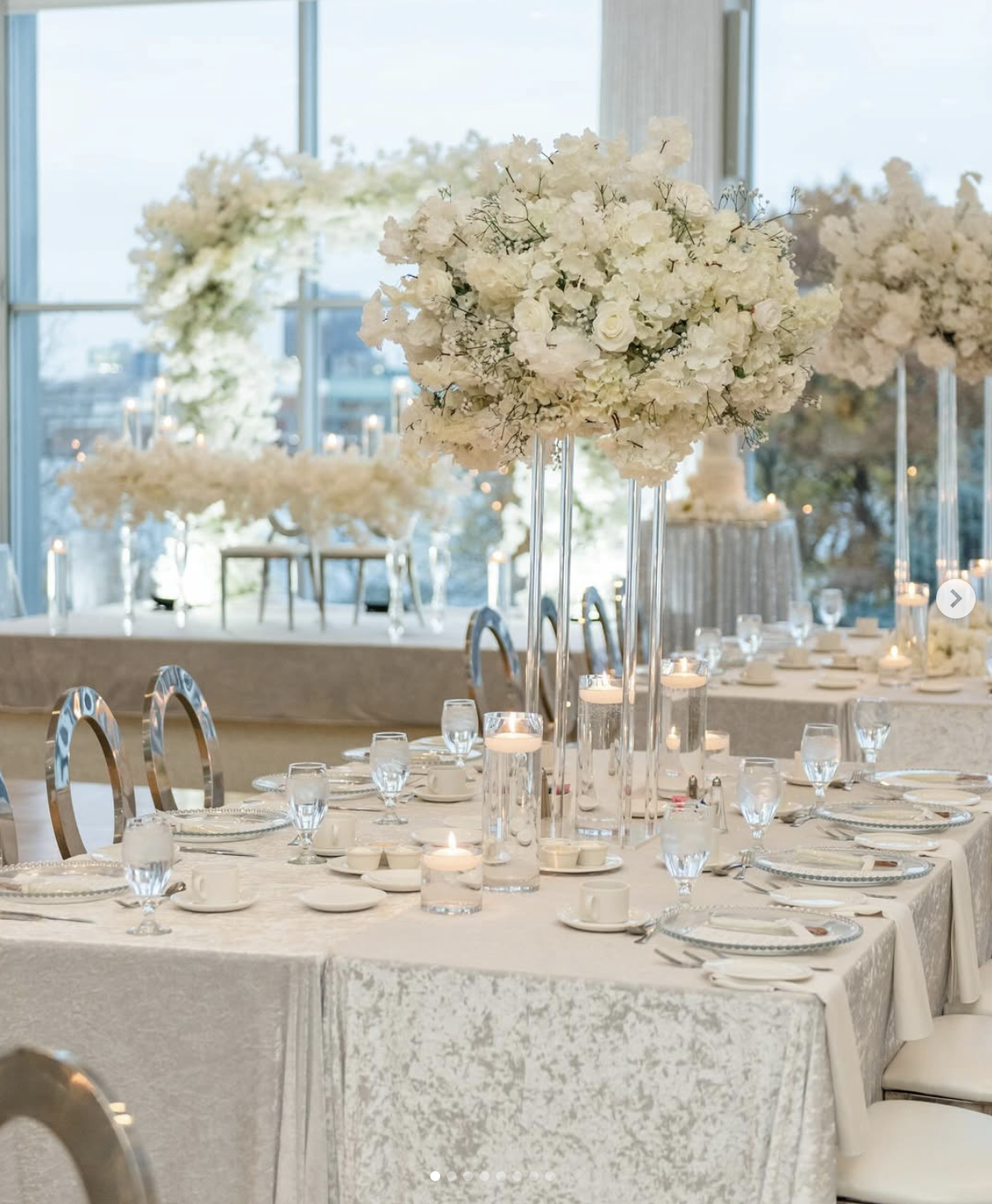 Elegant banquet table decorated with large floral centerpieces of white flowers, surrounded by glassware, white china, and candles, in a bright setting with large windows.