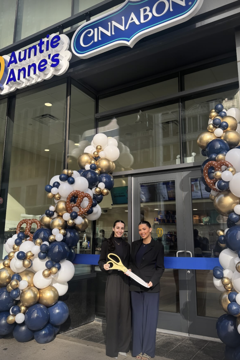 Two women standing in front of a Cinnabon and Auntie Anne's store with balloon decorations, holding large scissors for a grand opening or ribbon-cutting ceremony. Chicago, Millenium Park, Grant Park