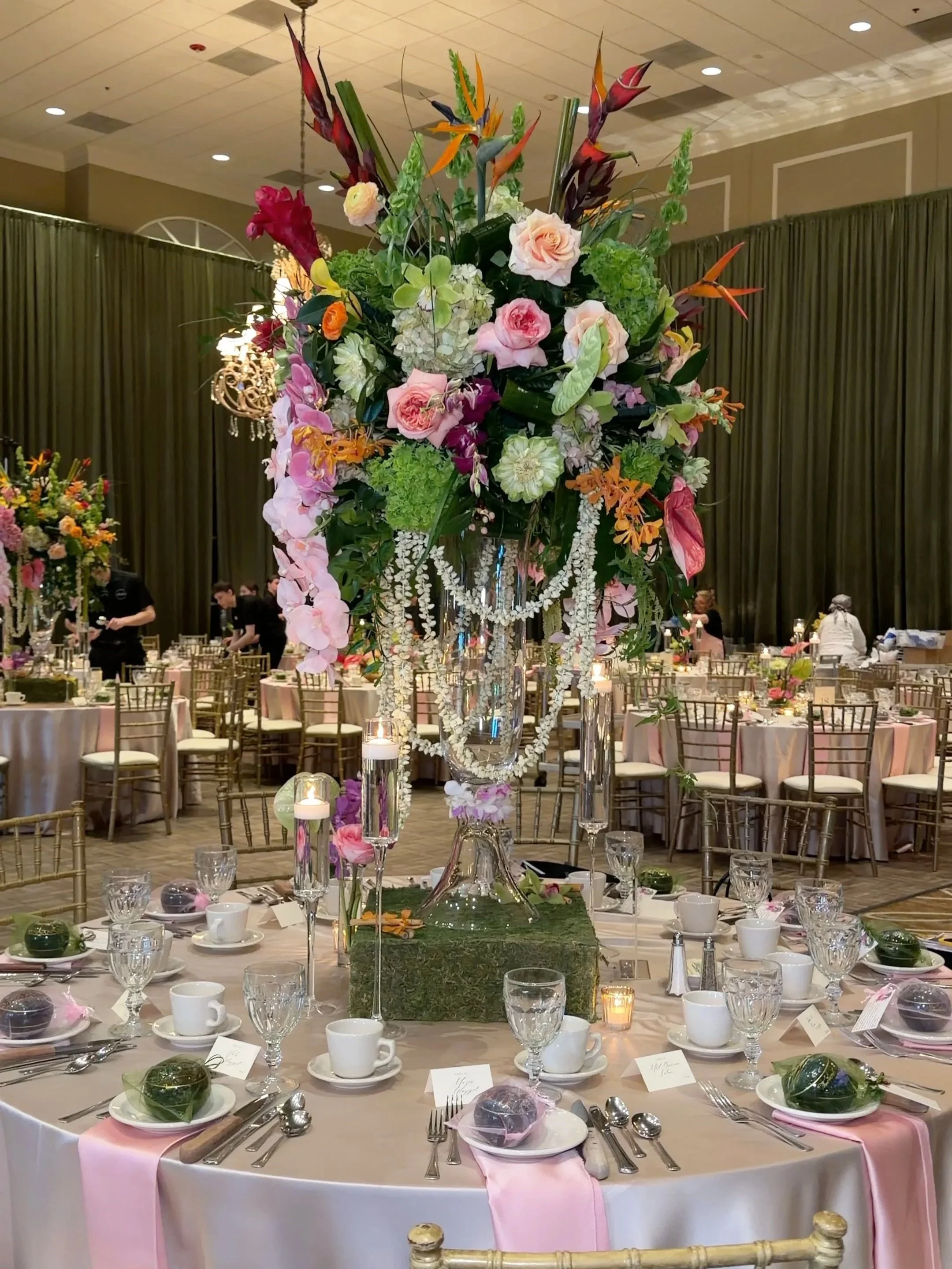 A large floral centerpiece on a tall glass vase with hanging pearl garlands is situated on a decorated banquet table at a formal event. The table is covered with a white tablecloth, set with white cups and plates, surrounded by gold chairs, and has a small candle in a glass holder. The floral arrangement includes roses, orchids, lilies, and other colorful flowers. The background shows more tables with floral centerpieces, a high ceiling, and green curtains.