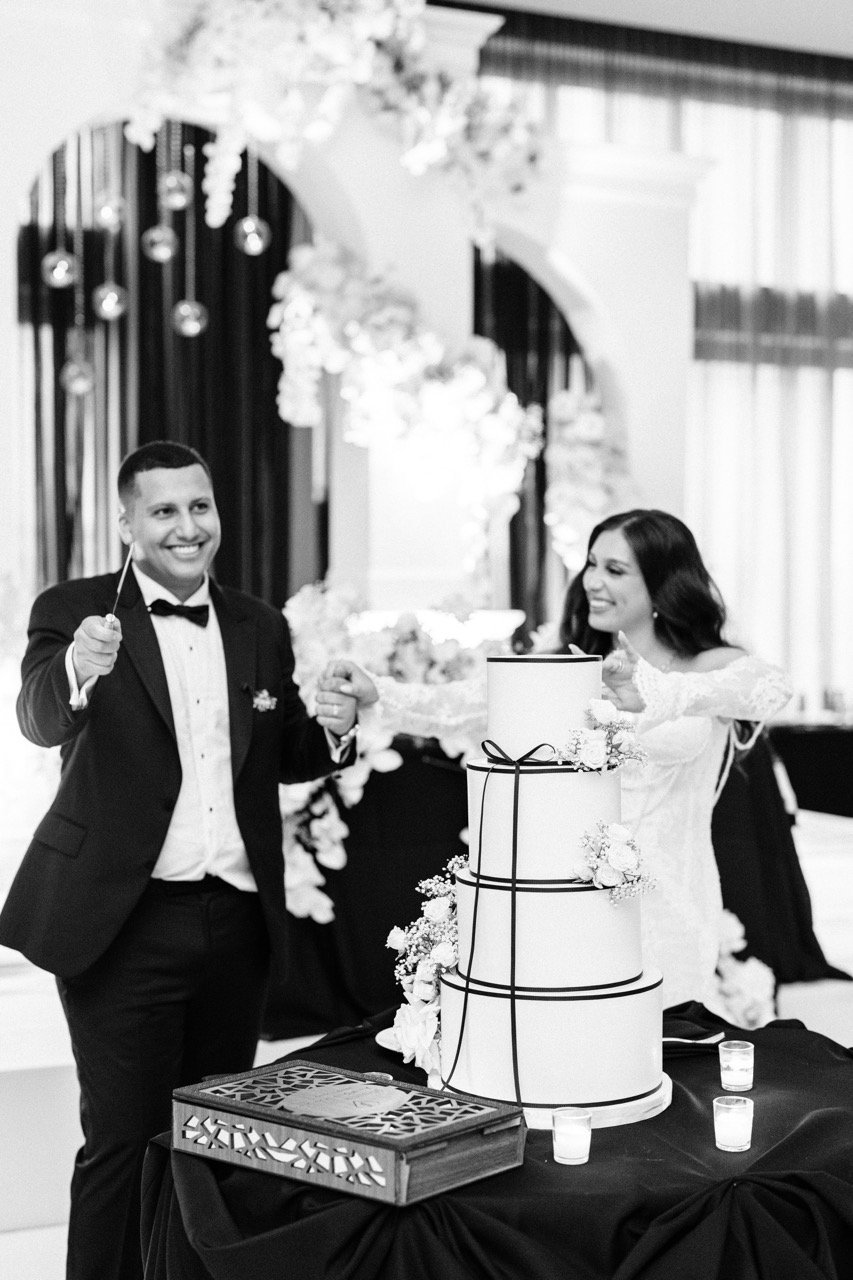 A black and white photo of a bride and groom cutting a wedding cake together. The groom is in a tuxedo, smiling, holding a knife, and the bride, in a wedding dress, is smiling and holding a knife. The cake is tall, tiered, and decorated with flowers. The background features floral arrangements and curtains.