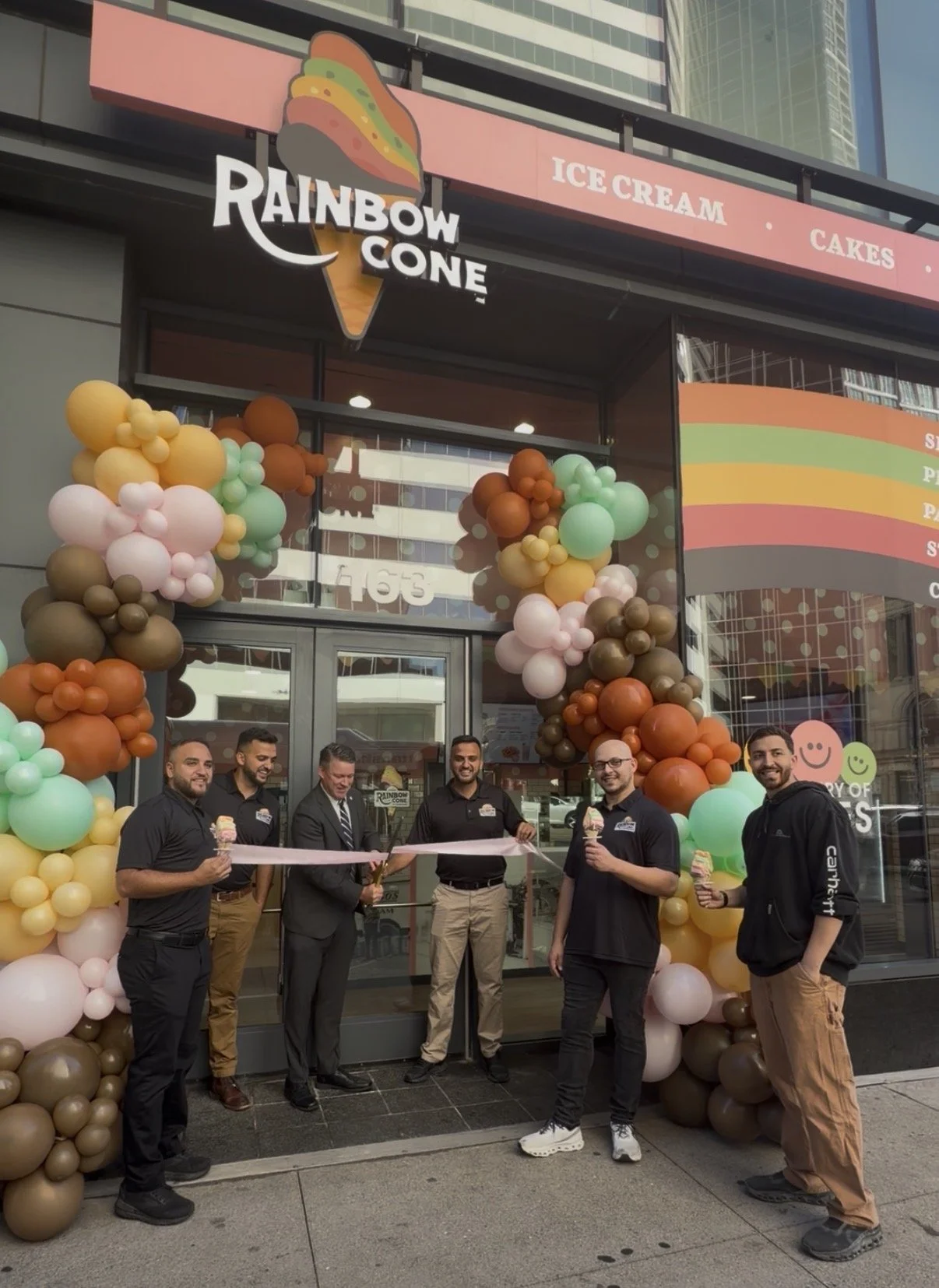 Group of five men standing in front of Rainbow Cone ice cream shop opening celebration, holding ice cream cones, surrounded by colorful balloon decorations, with a large ribbon being held for a ribbon-cutting ceremony.