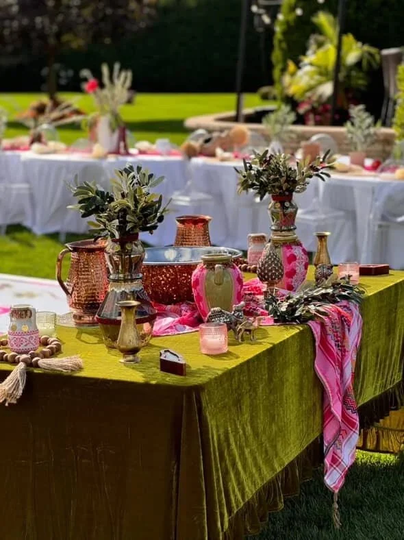 Decorative table setup outdoors with vases, flowers, candles, and small decorative items on an olive green tablecloth, with additional tables and greenery in the background.