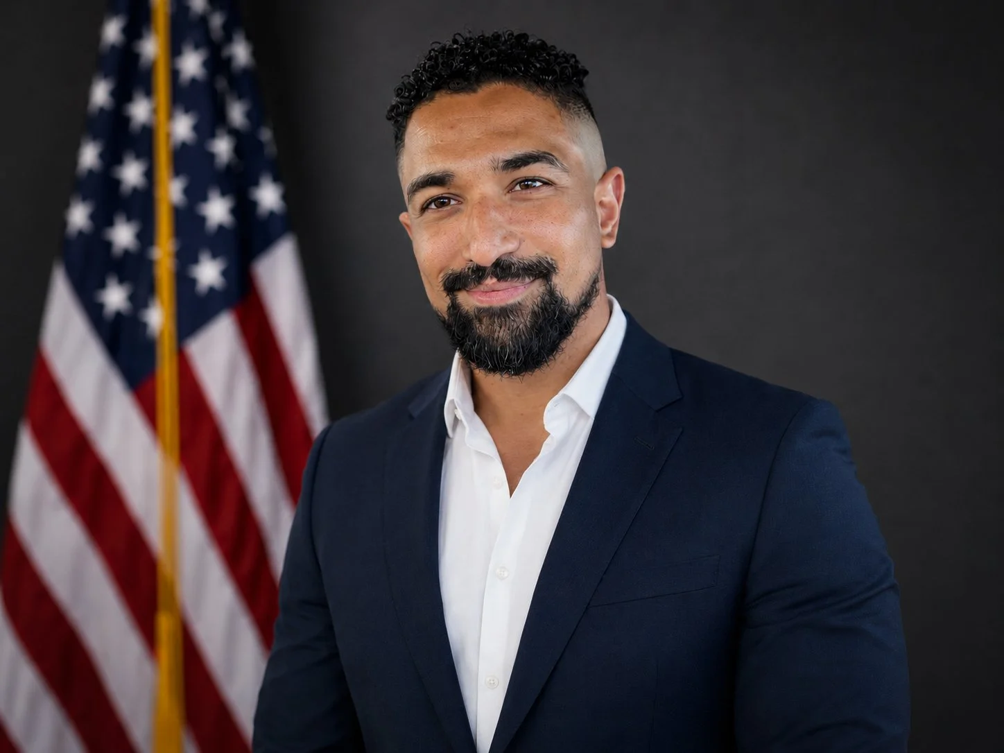 A man with short curly black hair, a beard, and wearing a dark suit with a white shirt. Behind him is a blurred American flag on a dark background.