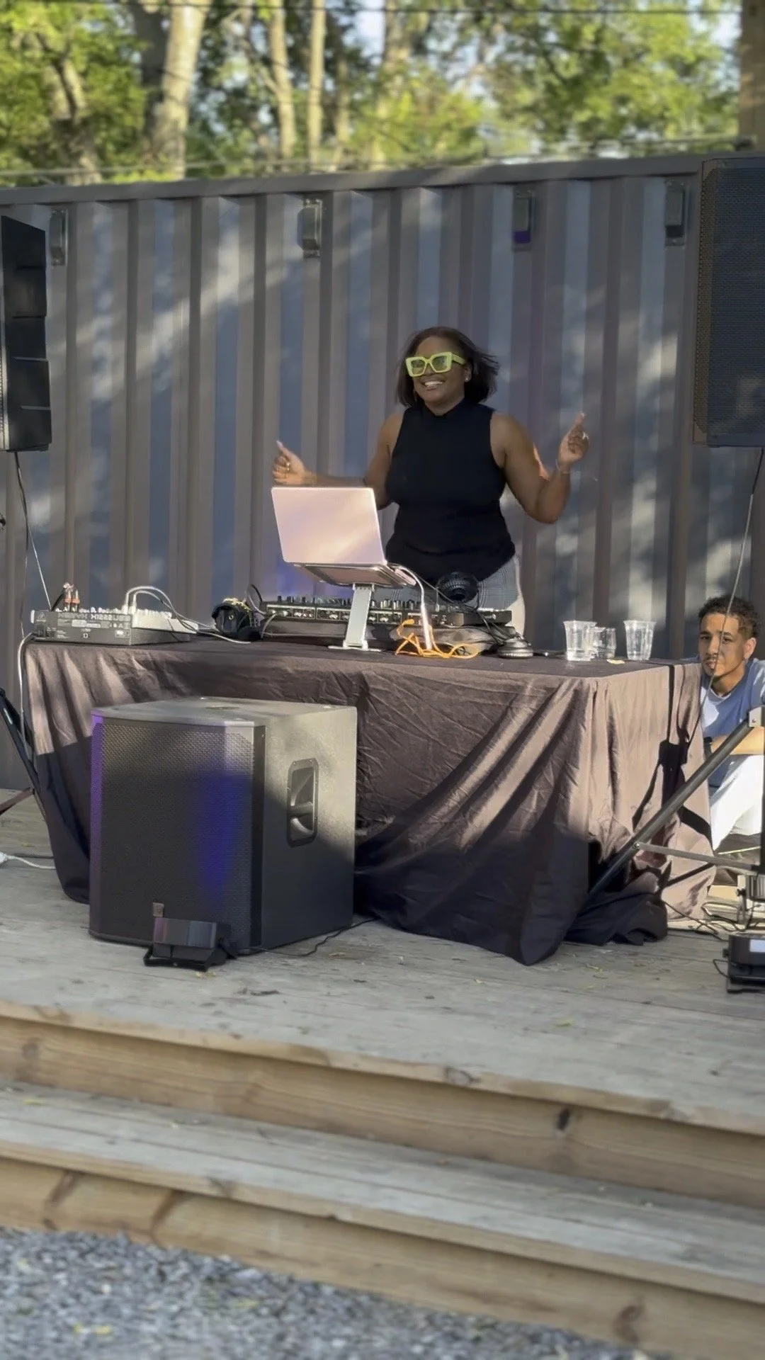 A woman DJ wearing yellow sunglasses and a black sleeveless top, standing behind a DJ setup with speakers, laptop, and equipment, outdoors in front of a metallic wall, with a young person sitting nearby.