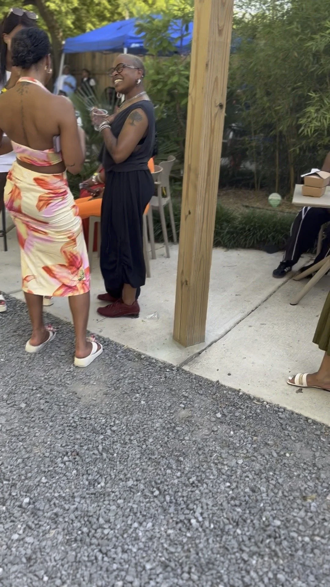 Two women standing and conversing outdoors during daytime at a social gathering. One woman is wearing a floral dress and white sandals, the other in a black dress with red shoes. There are people in the background, some standing and some sitting, und