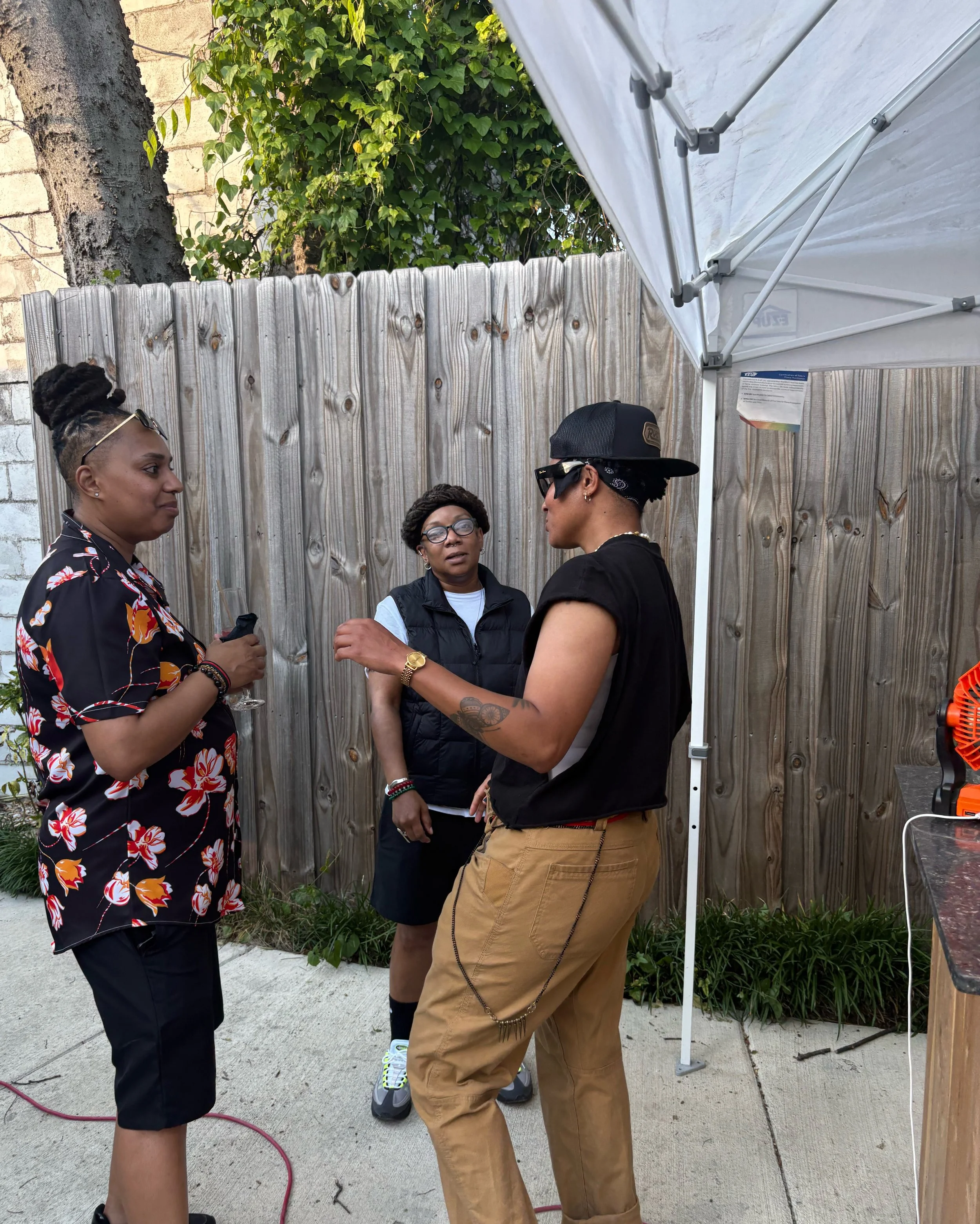 Four people standing in a semi-circle outside, having a conversation near a wooden fence and a canopy tent. Three women and one man, some holding glasses, appear to be engaged in a group discussion.