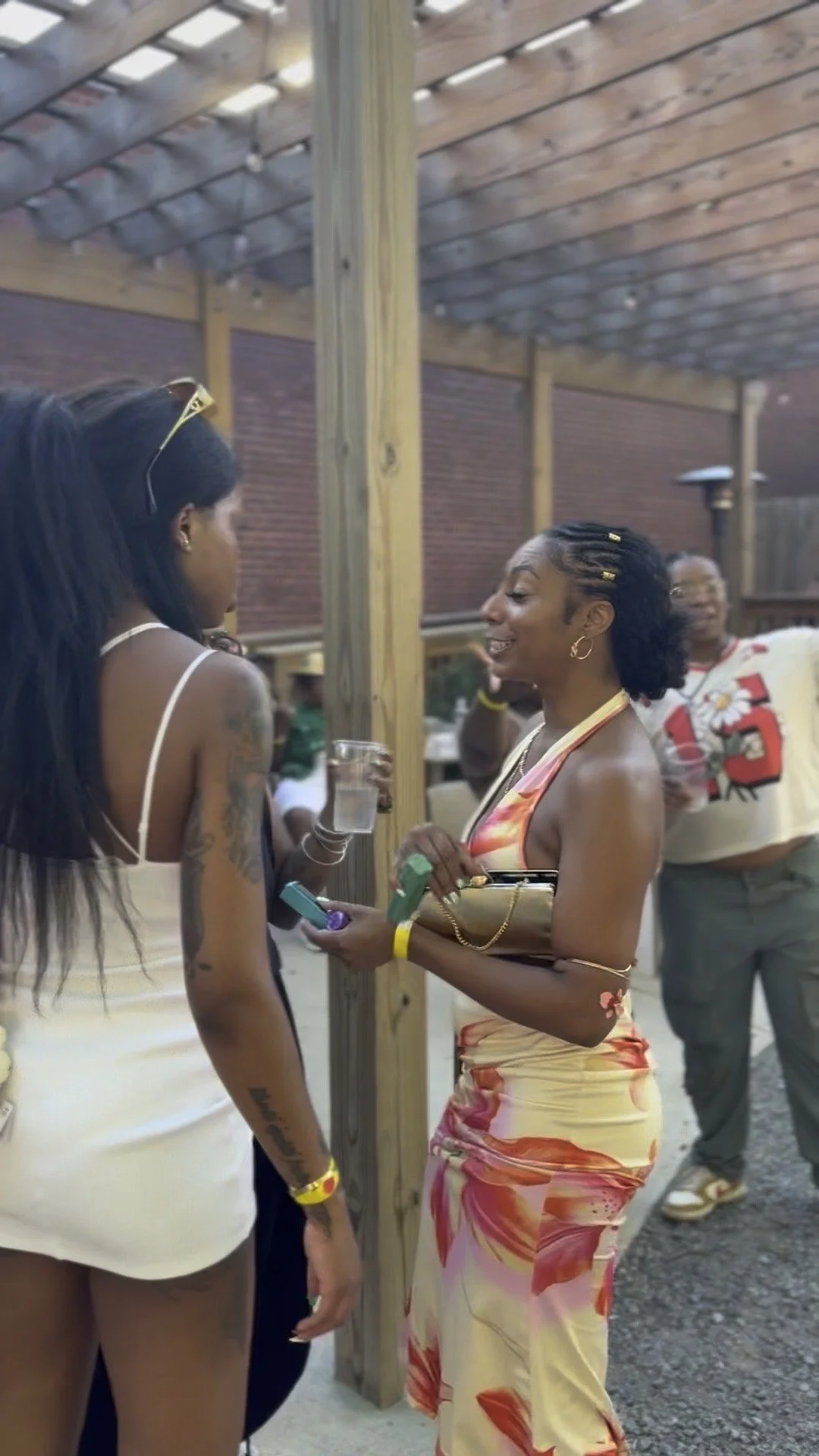 Two women having a conversation at an outdoor gathering. One woman with long dark hair and tattoos wearing a white dress, and another woman with short curly hair in a colorful floral dress. They are smiling and holding drinks and small items in their