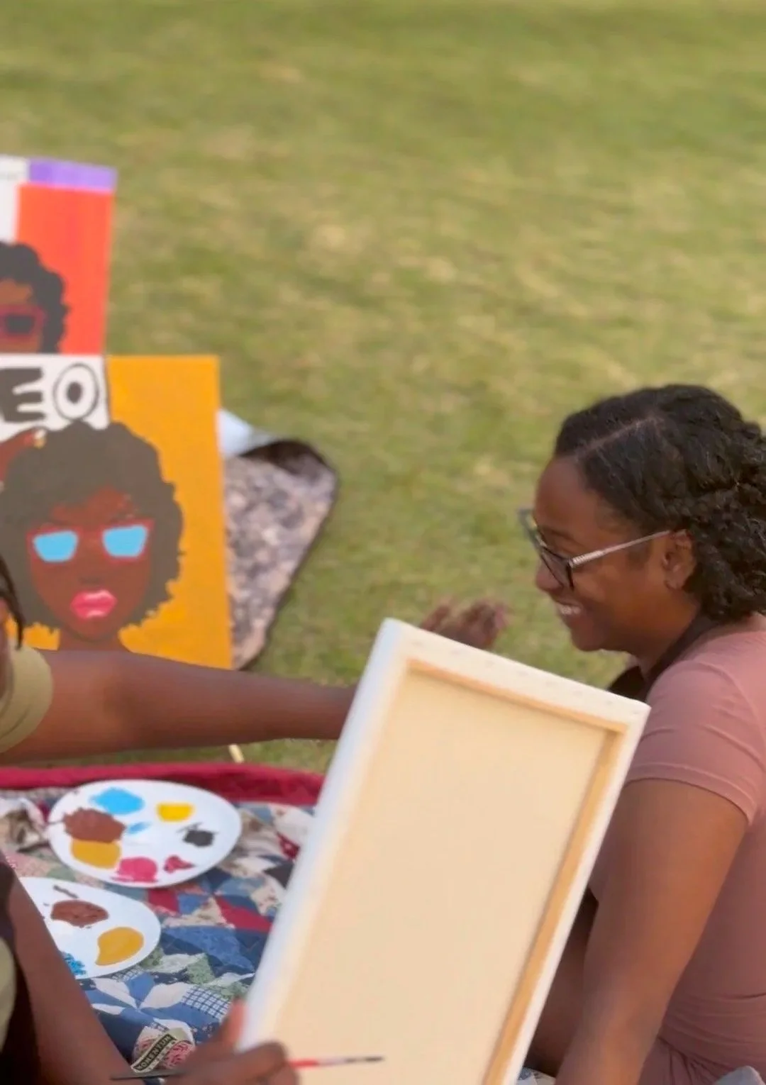 Two women smiling, one sitting at an outdoor art event with painting supplies, and artwork featuring a woman with glasses and textured hair on display.