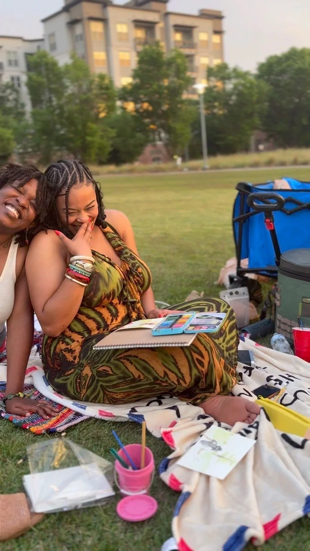 Two women sitting on a blanket in a park, laughing and smiling, with art supplies and backpacks around them, and a large building and trees in the background.