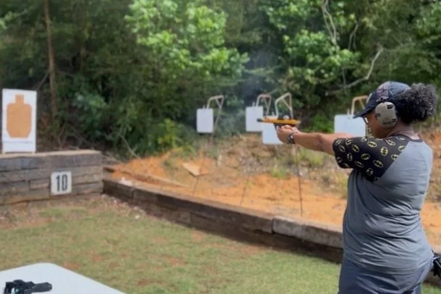 A woman shooting a handgun at outdoor shooting targets at a gun range. She is wearing safety headphones, a baseball cap, and a t-shirt with a Batman logo pattern, aiming the gun downrange.