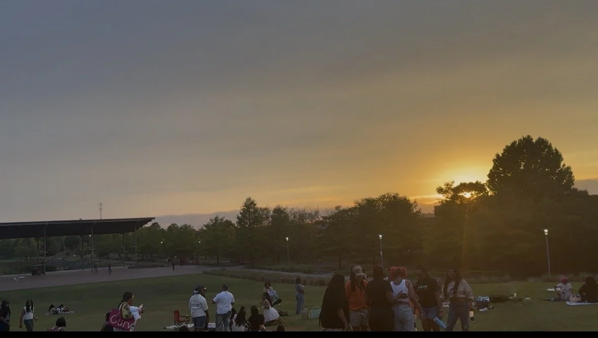 People gathered outdoors in a park during sunset, some standing and some sitting on the grass, with trees and a large shelter structure in the background.