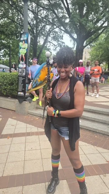 A smiling woman with curly hair stands outdoors with a rainbow umbrella, wearing rainbow-striped knee-high socks, a black sleeveless top, denim shorts, and multiple bracelets. She is holding a rainbow umbrella and has a lively group of people in the 