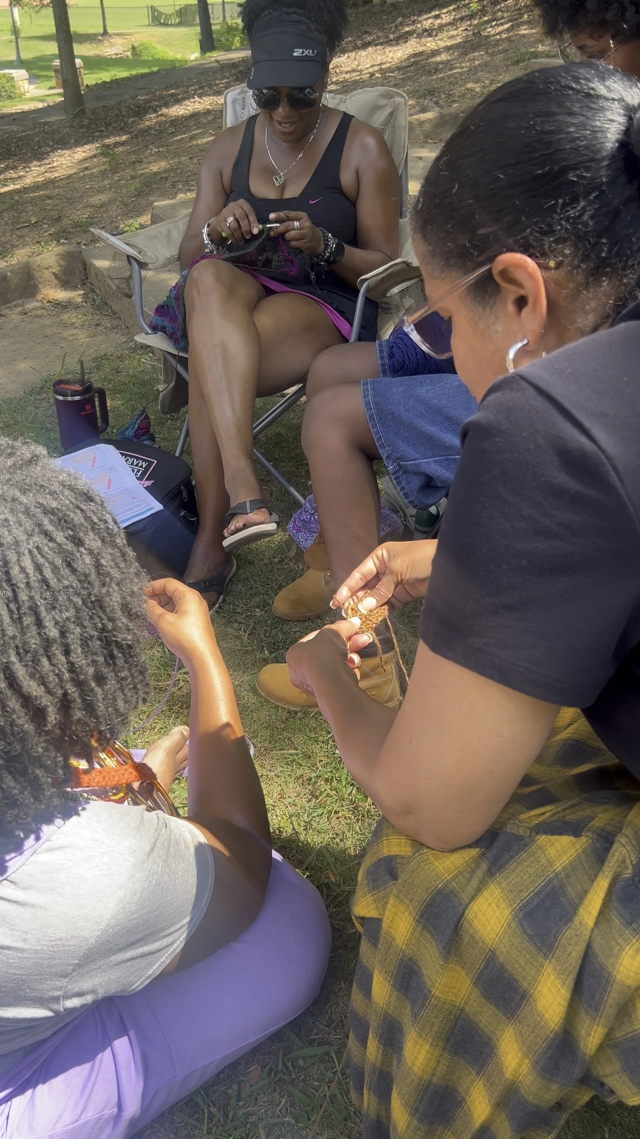 A group of women and children sitting outdoors in a park, engaged in knitting or crafting, with trees and grass in the background.