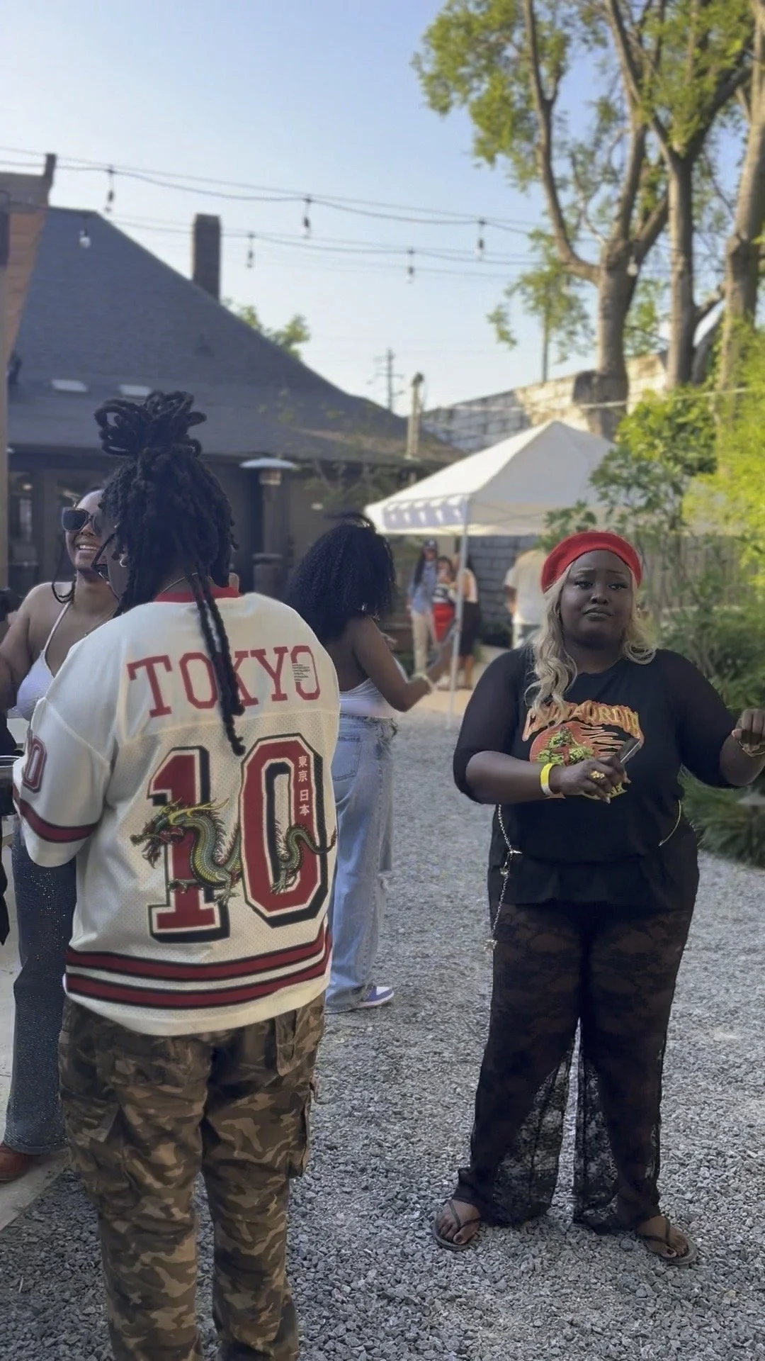 A group of diverse people socializing outdoors during a sunny day, with a woman in the foreground wearing a red headband, black and red 'Harley-Davidson' T-shirt, and lace pants, holding a drink, while others are smiling and chatting in the backgroun