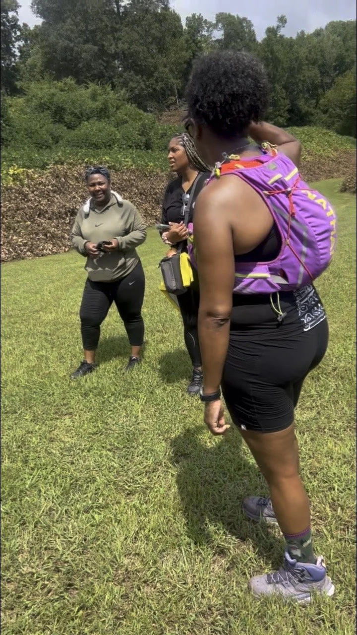 Four women dressed in athletic gear, standing on grass in a park-like area with trees in the background, engaged in conversation during a hike or outdoor activity.