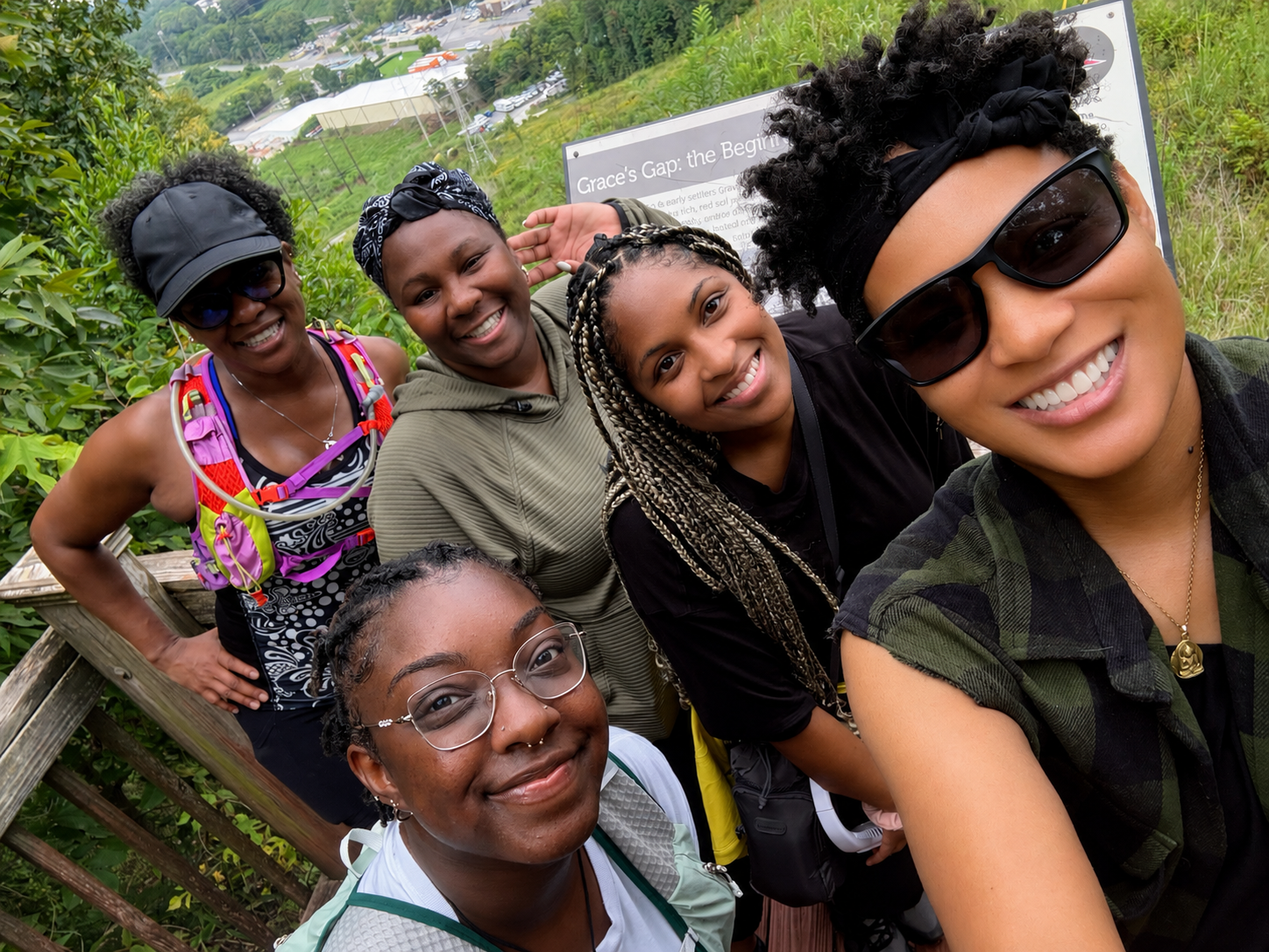 Six smiling women posing for a selfie outdoors on a wooden platform, with greenery and a sign in the background.
