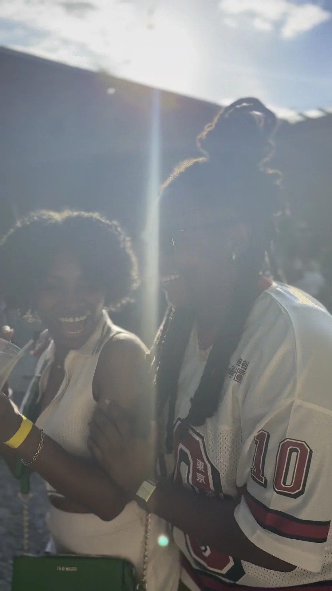Two people smiling and talking outdoors in sunlight, one wearing a white shirt and the other wearing a sports jersey with red and black accents.