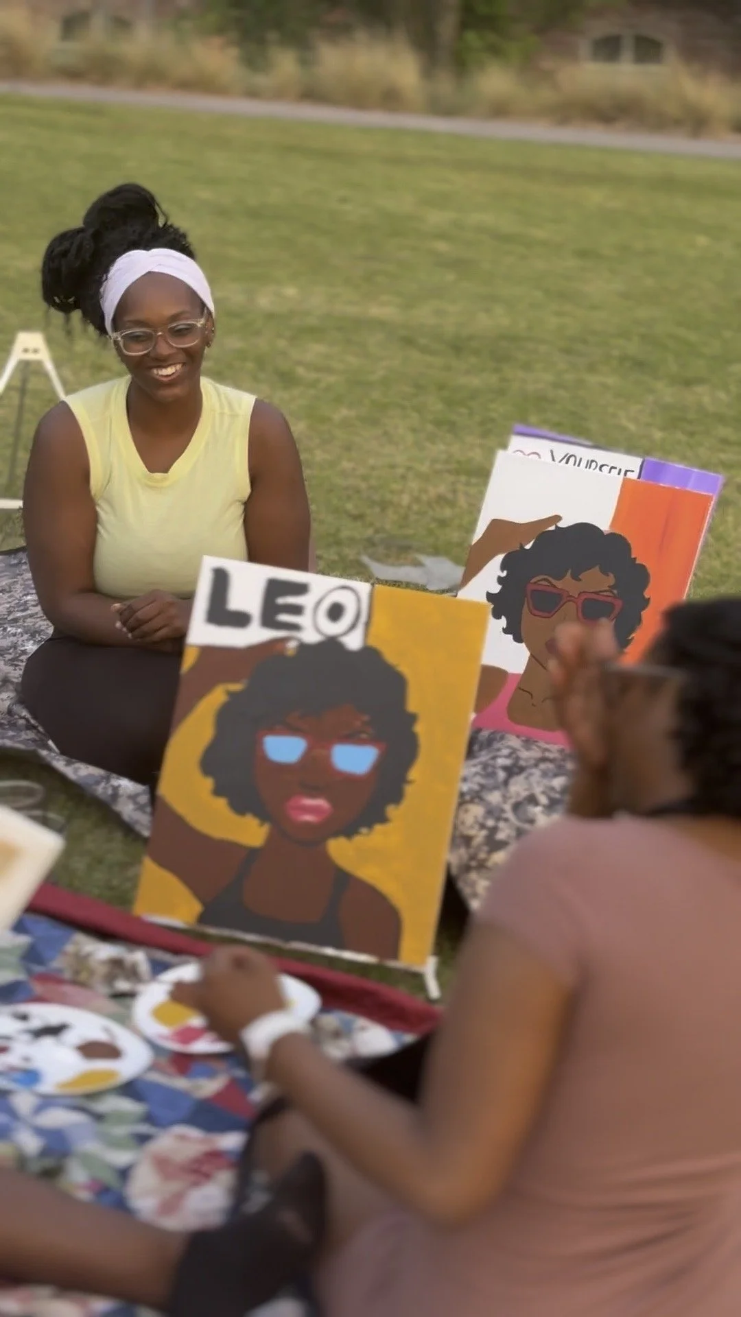A woman sitting on a blanket outdoors at a table with art supplies, smiling at children displaying colorful illustrations of a woman with curly hair and sunglasses.