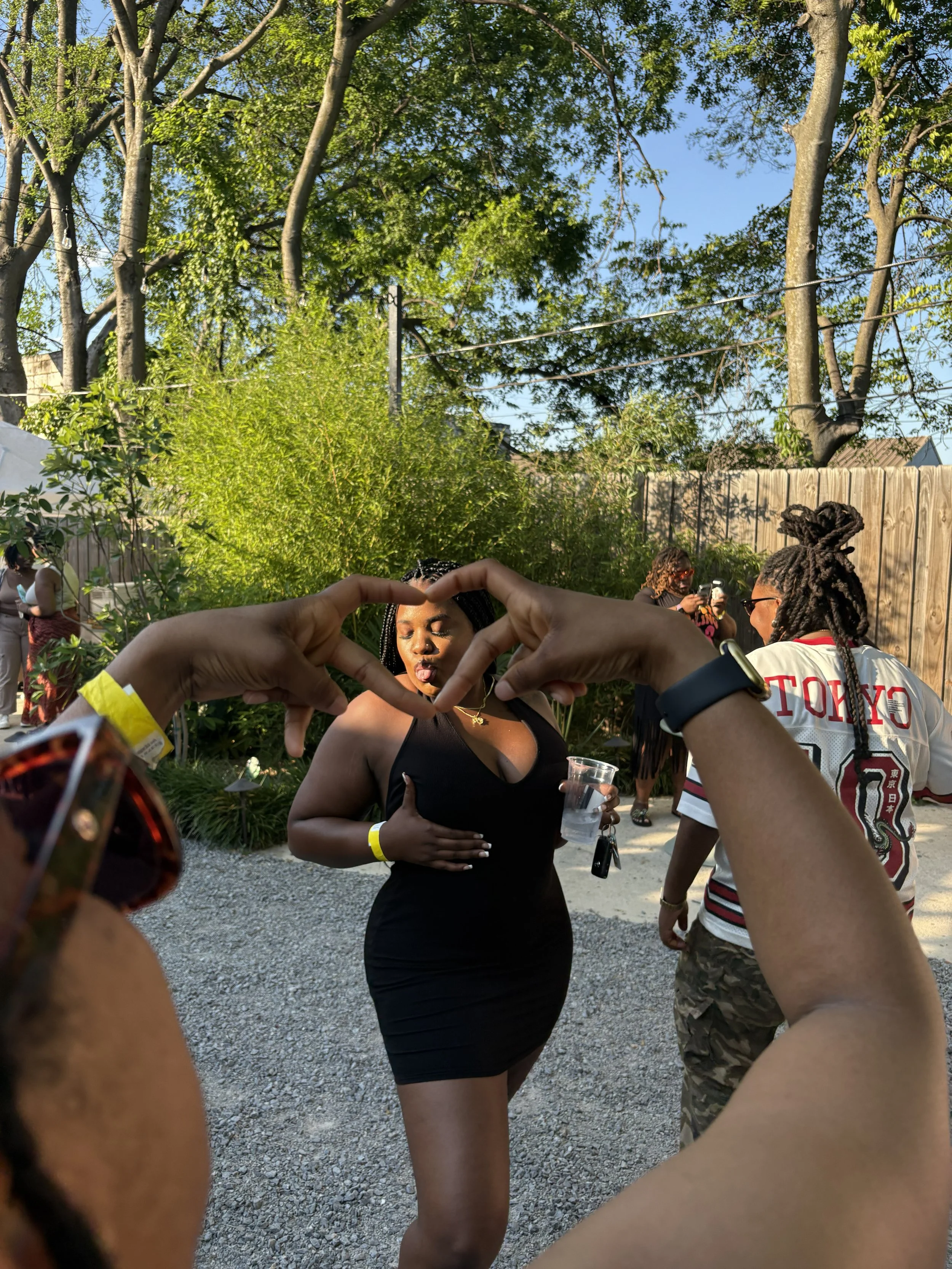 A woman in a black dress making a heart shape with her hands at an outdoor gathering with trees and other people in the background.