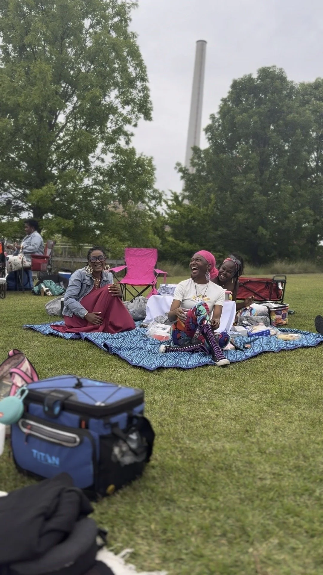 Four women sitting on a blanket in a park, laughing and enjoying each other's company; picnic items and chairs around them, with trees and a tall smokestack in the background.